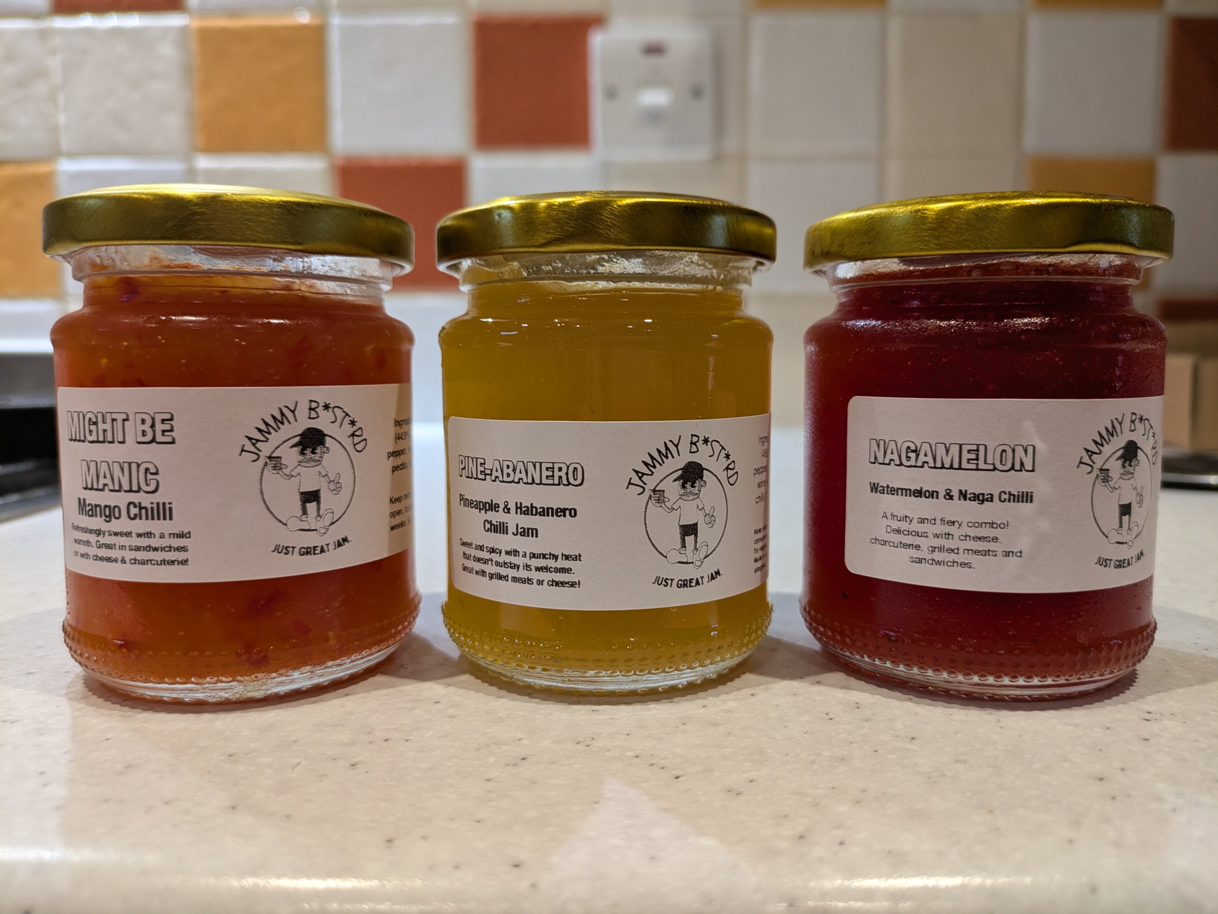 Three jars of jam with gold-colored lids on a kitchen countertop, with a colorful tiled backsplash in the background.