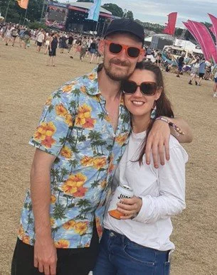 A man and woman smiling outdoors at a festival, with the man wearing a colorful Hawaiian shirt, sunglasses, and a hat, and the woman wearing sunglasses and holding a beverage.