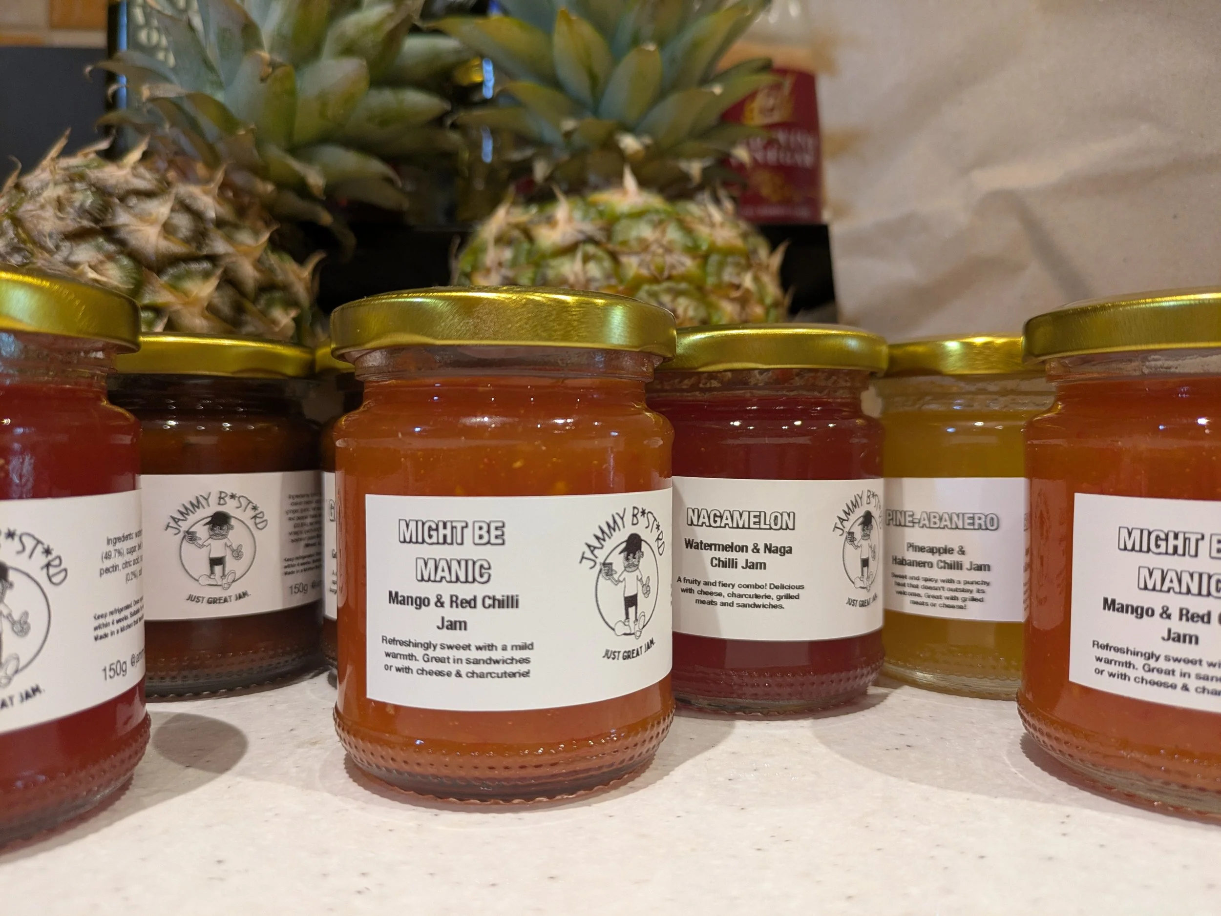 Several jars of jam labeled with various flavors, such as Mango & Red Chilli, Watermelon & Naga Chilli, Pineapple & Habanero, and Mango & Red Chilli, with pineapples in the background.