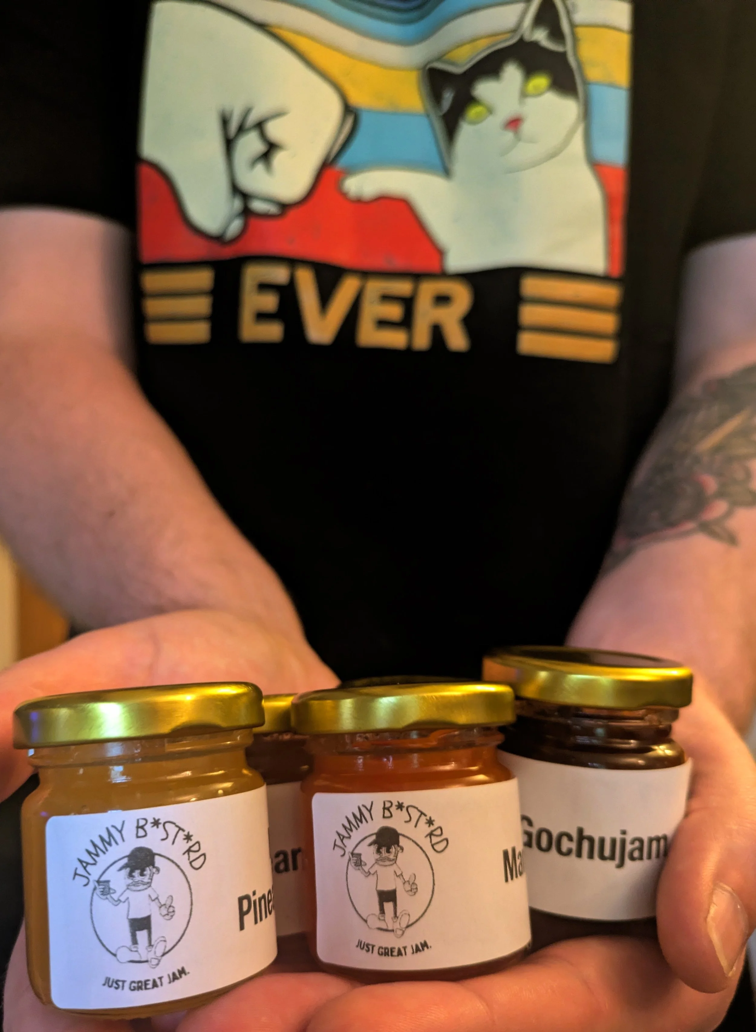 A person holding jars of jam with white labels and gold caps. The labels have a logo with a man holding a jam jar and the text 'JAMMY B*ST*RD' and 'JUST GREAT JAM'.