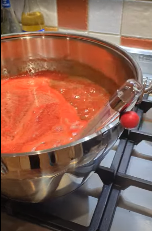 A stainless steel pot on a stove filled with boiling red liquid as part of the jam making process.