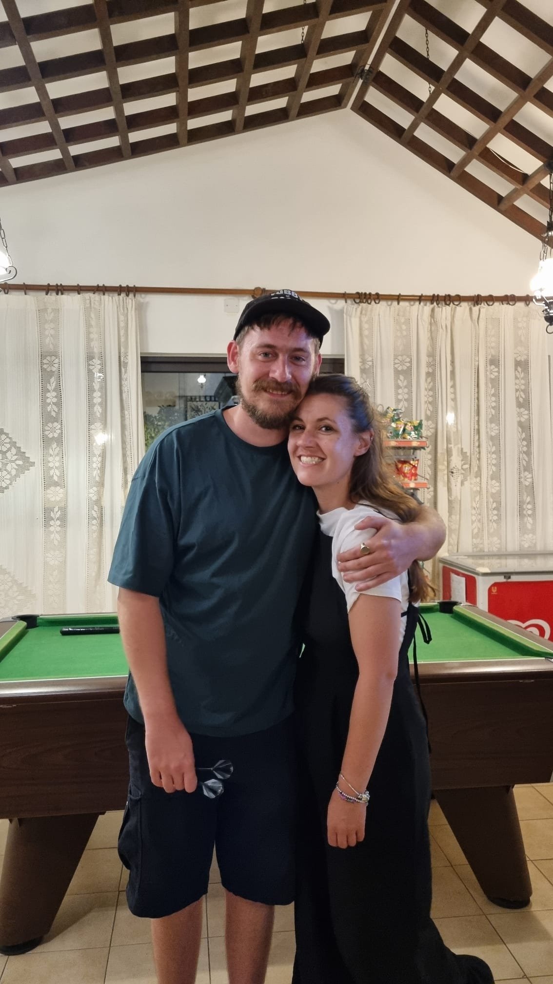 A man and woman hugging and smiling in front of a pool table in a cozy room with lace curtains and holiday decorations.