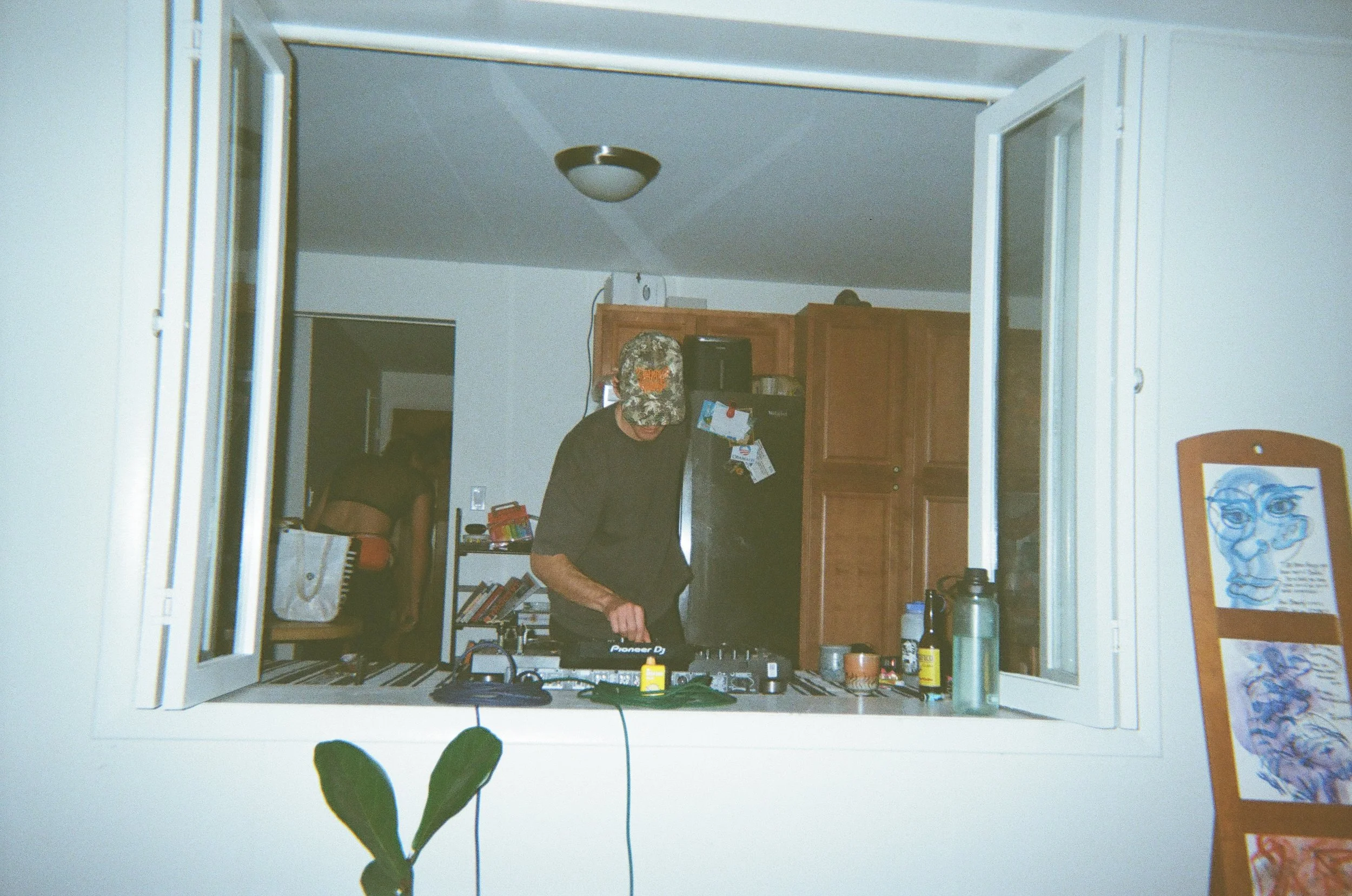 A person wearing a camouflage hat is standing behind a DJ controller, inside a room visible through a fireplace. The room contains a wooden cabinet, various items on the counter, and a picture of a cartoon character with glasses on the right side.