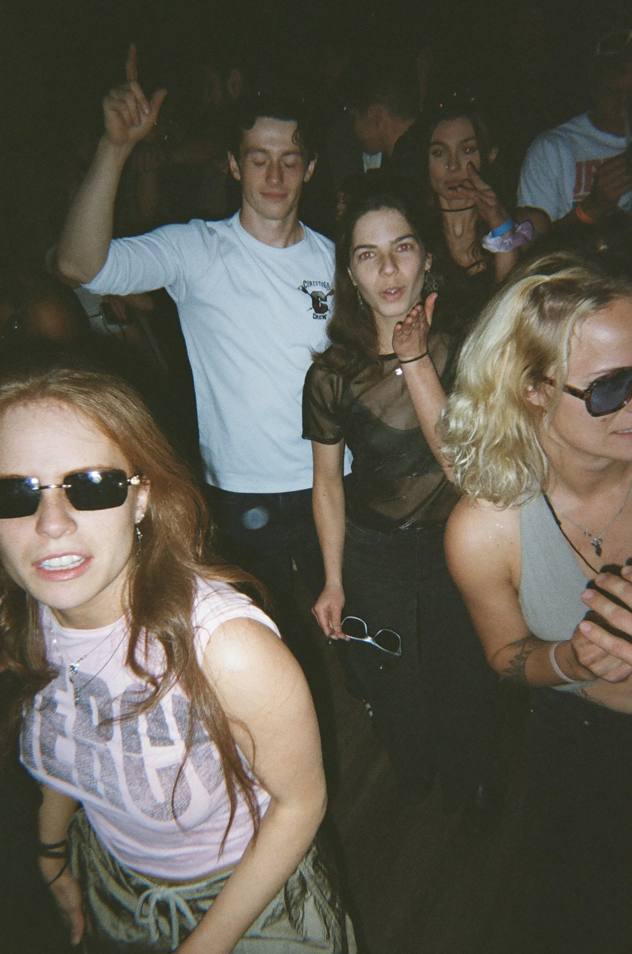 A group of people at a party taking a selfie. The person in the foreground has long red hair and is wearing sunglasses. Behind her, a woman with dark hair and a sheer black top, and a man with dark hair, wearing a white t-shirt, are posing for the pi