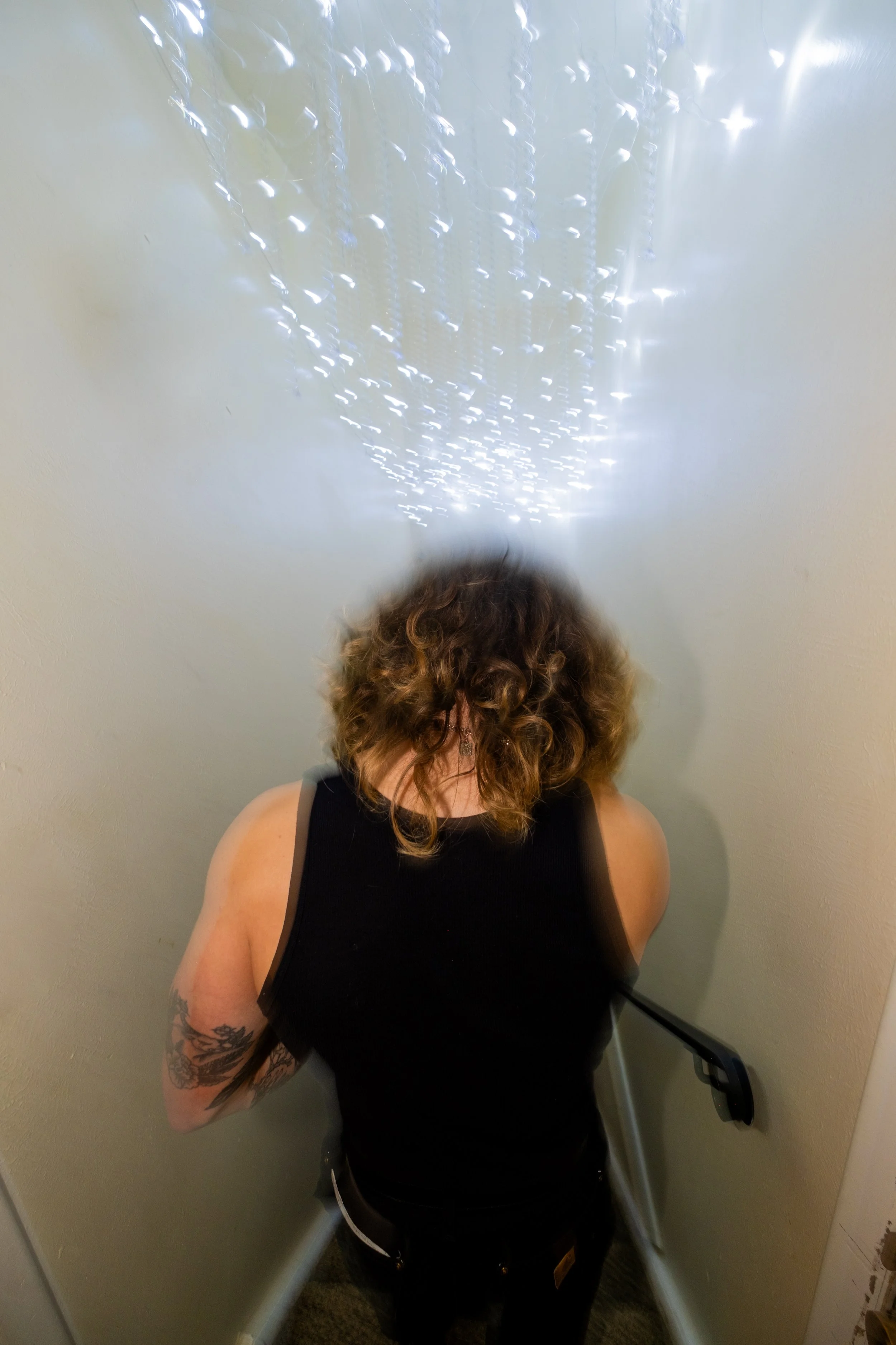 Person with curly hair, wearing a black sleeveless top, standing in an elevator, facing away from the camera.