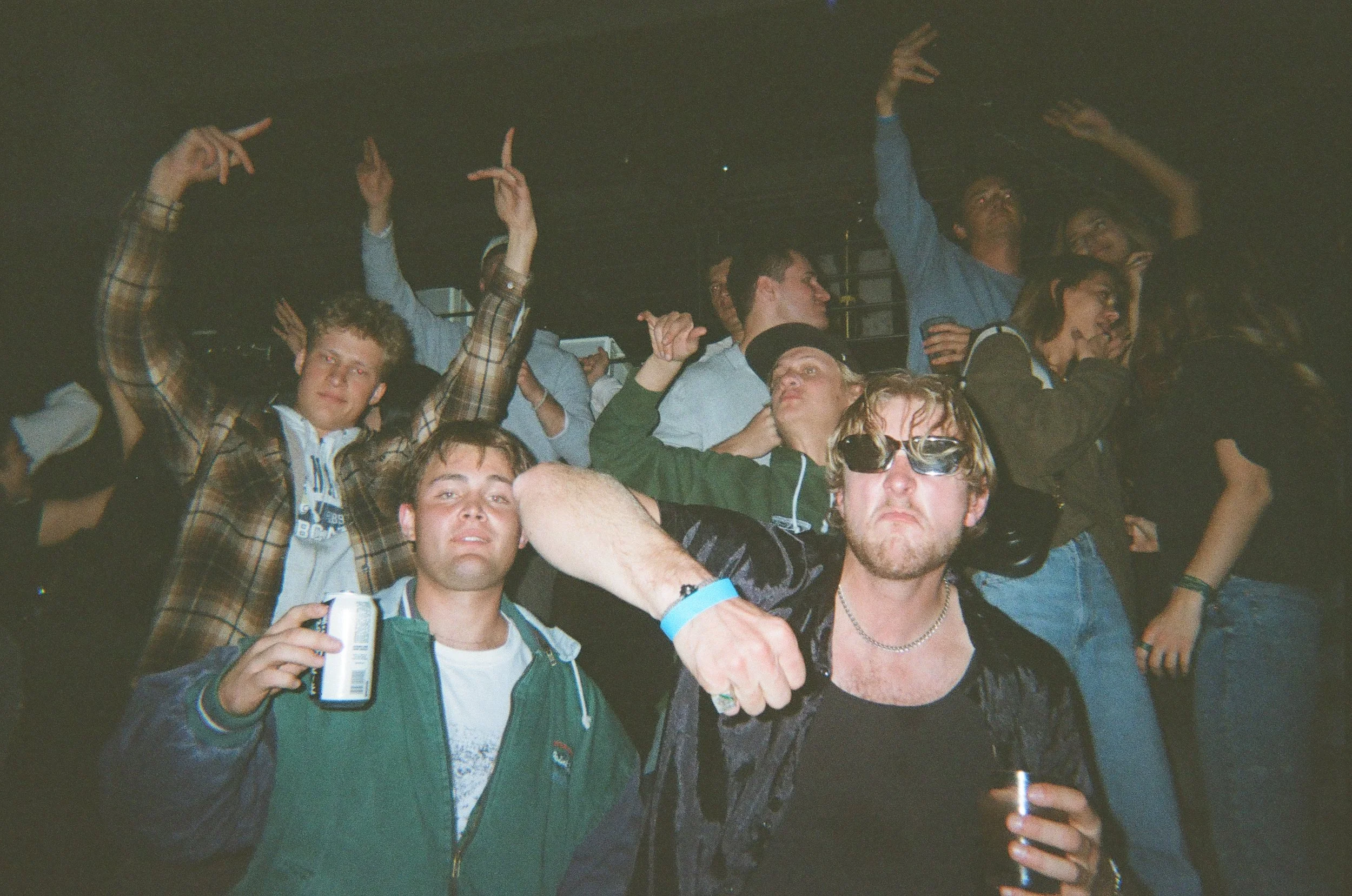 Group of young people dancing and cheering at a party or concert, some holding drinks, with a dark background.