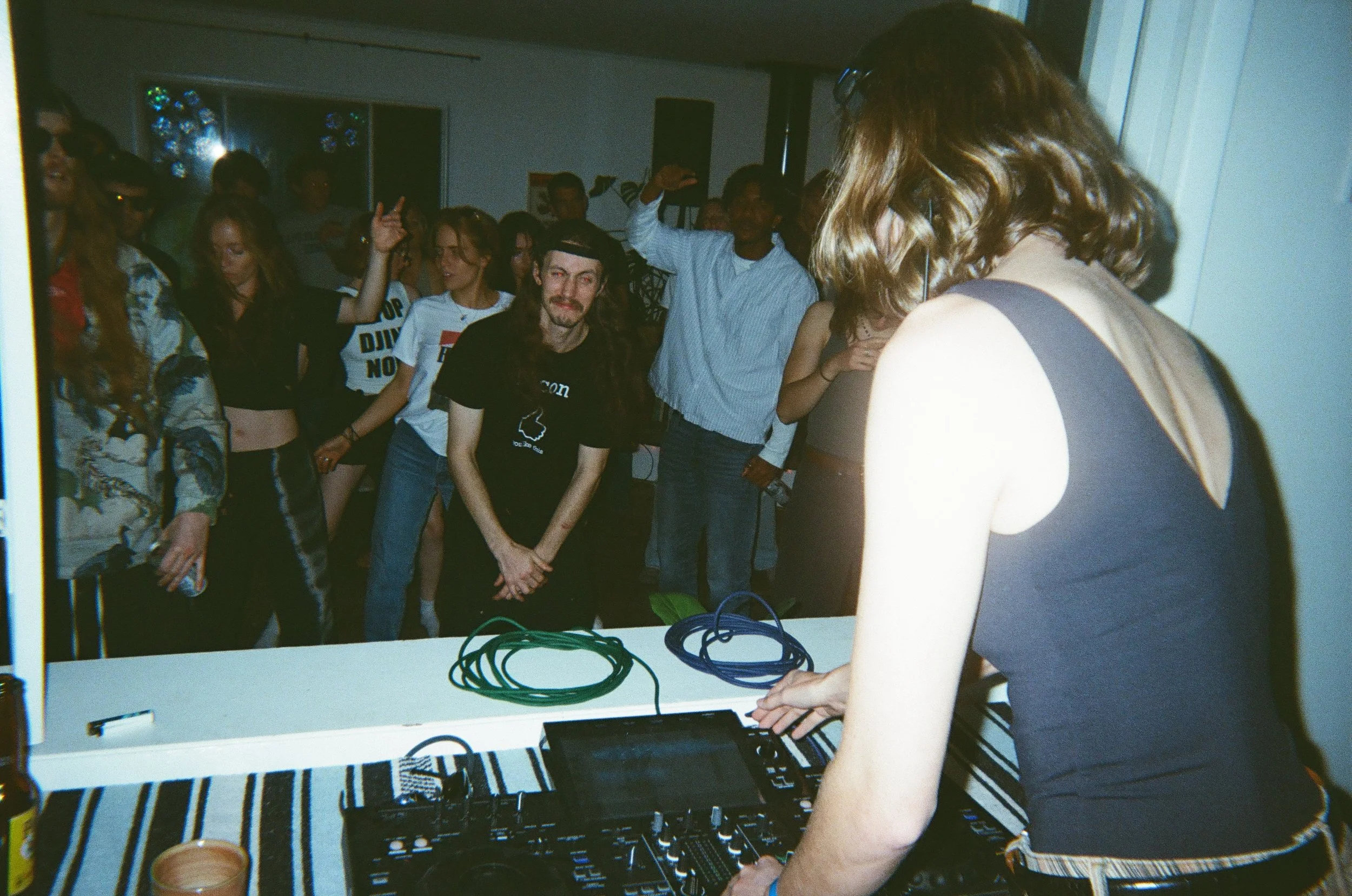 A DJ setup with a person in the foreground adjusting equipment, and a crowd of young people dancing and socializing at a party or club.