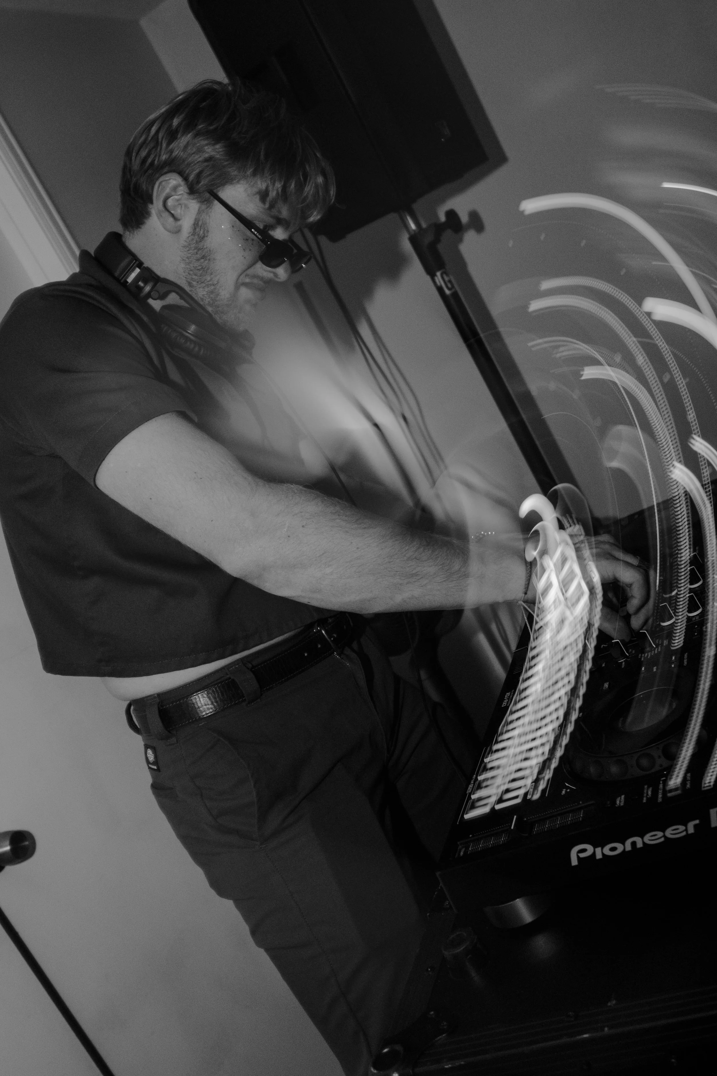 A DJ wearing glasses and headphones stands behind a Pioneer DJ equipment, adjusting the controls during a performance.