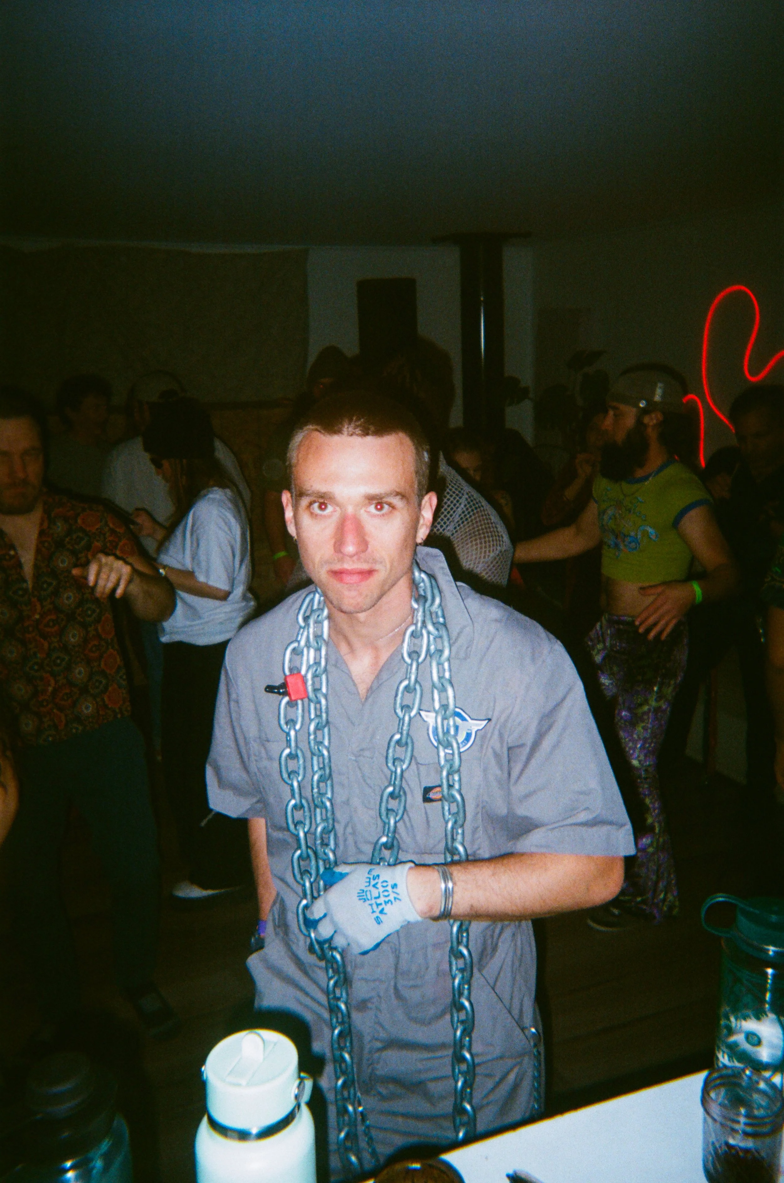 A young man wearing a gray shirt, white gloves, and large chain necklaces stands in a crowded room with a party or photo event atmosphere, with people in colorful casual clothing around him and various items on the table in front of him.