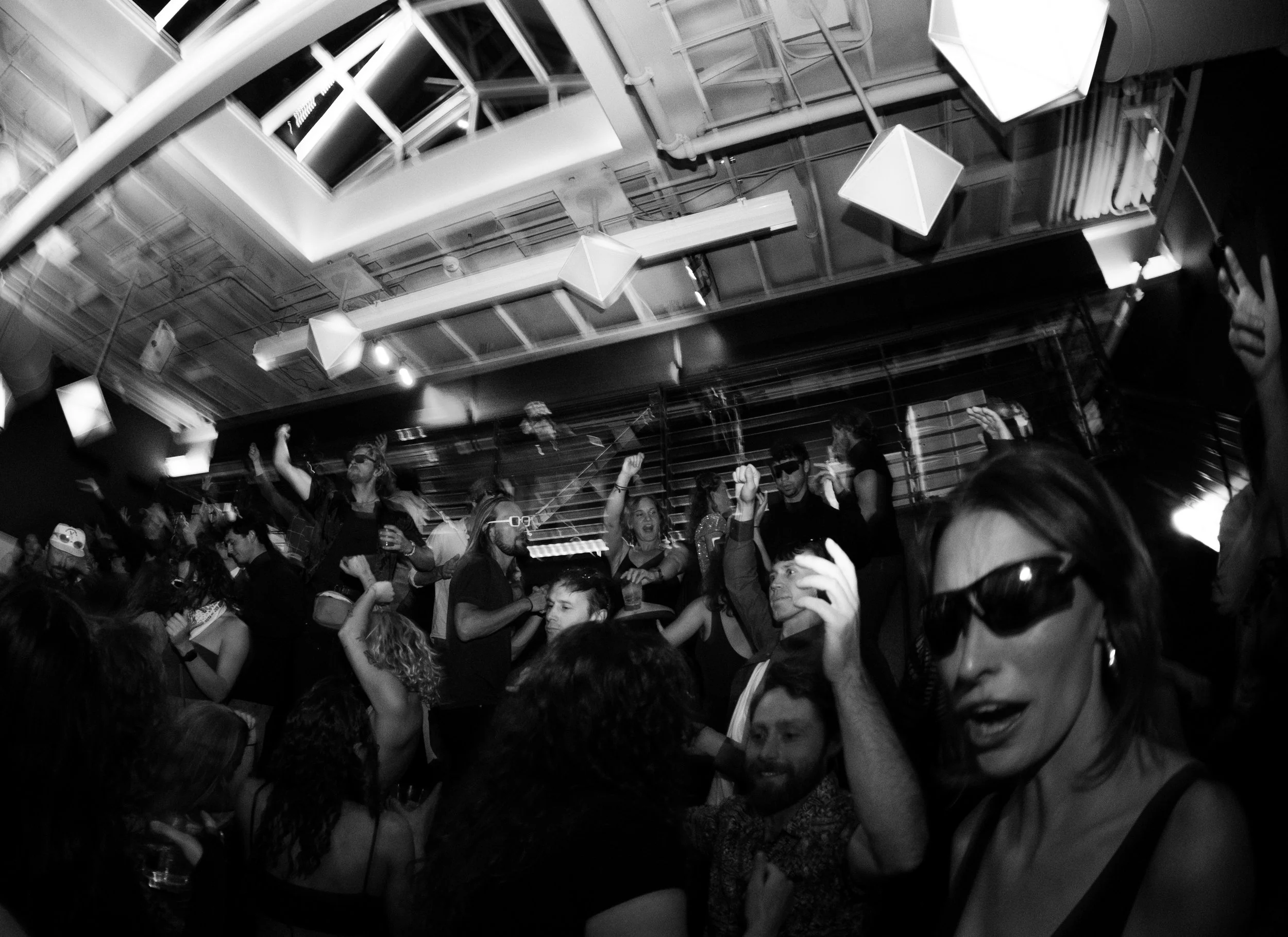 A crowd of people at a party or club, dancing and celebrating under modern ceiling lights in a dark room.