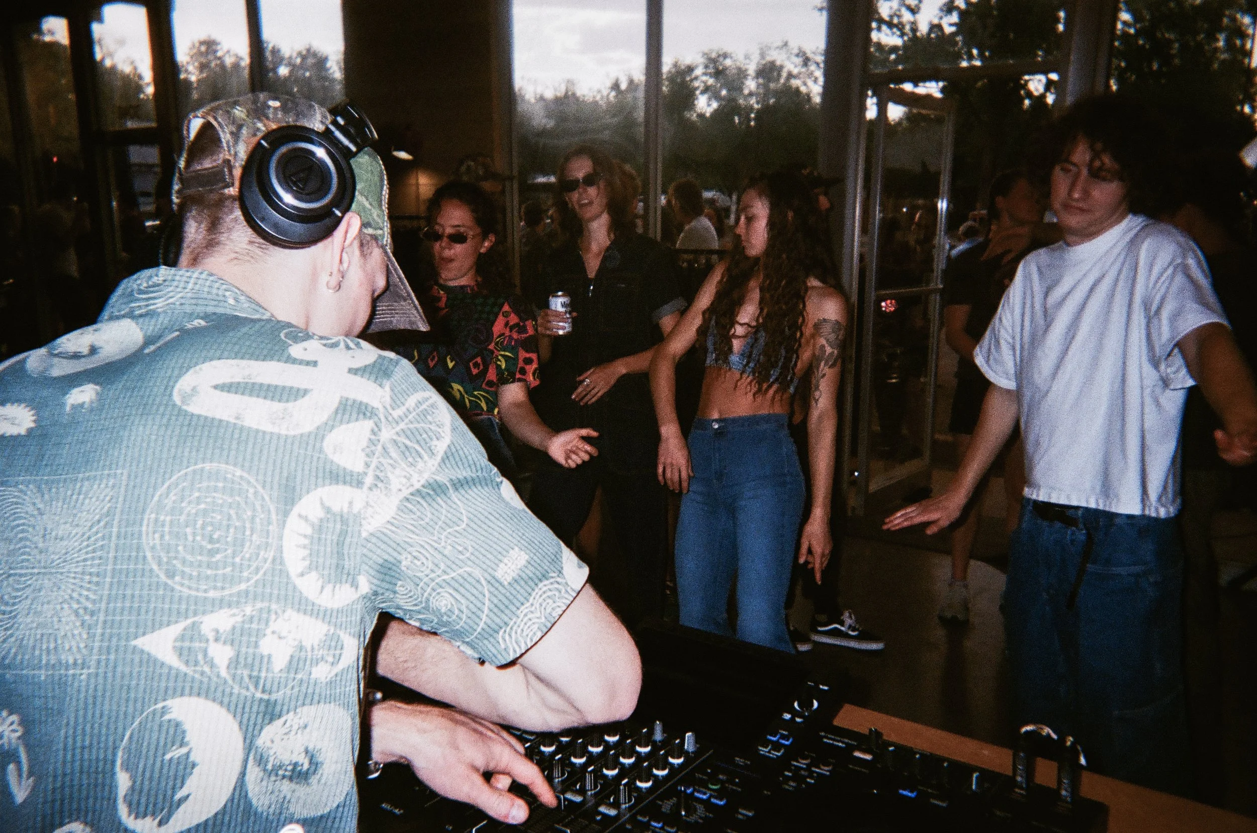 Person with large curly hair DJing at an indoor event, wearing a white T-shirt, standing behind a DJ mixer. Several people are dancing and socializing in the background near large windows showing outdoor trees.
