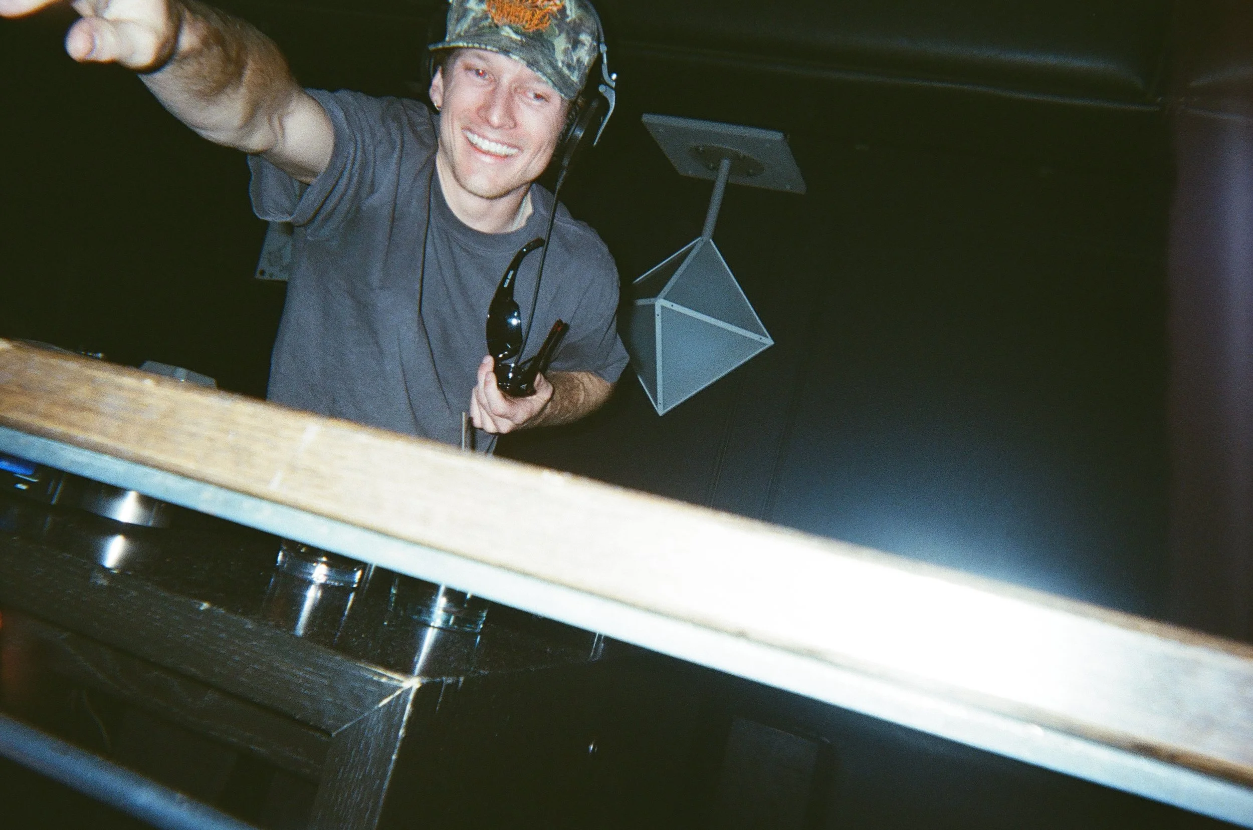 A smiling man wearing a camouflage hat and dark t-shirt, with headphones around his neck, pointing towards the camera. Boulder DJ
