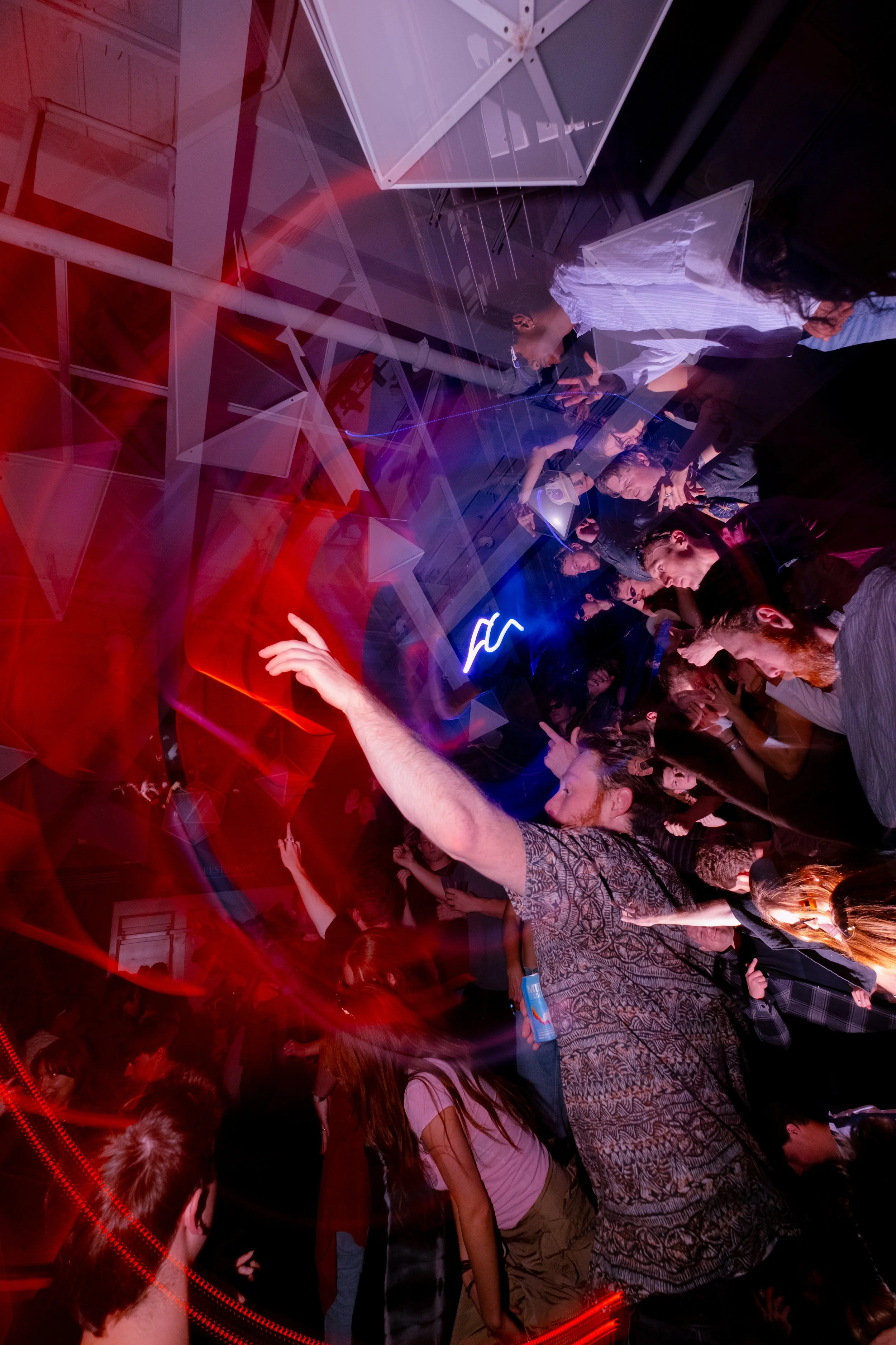 A crowded indoor party with people dancing and talking, illuminated by colorful red and blue lights, with reflective surfaces creating a vibrant atmosphere.