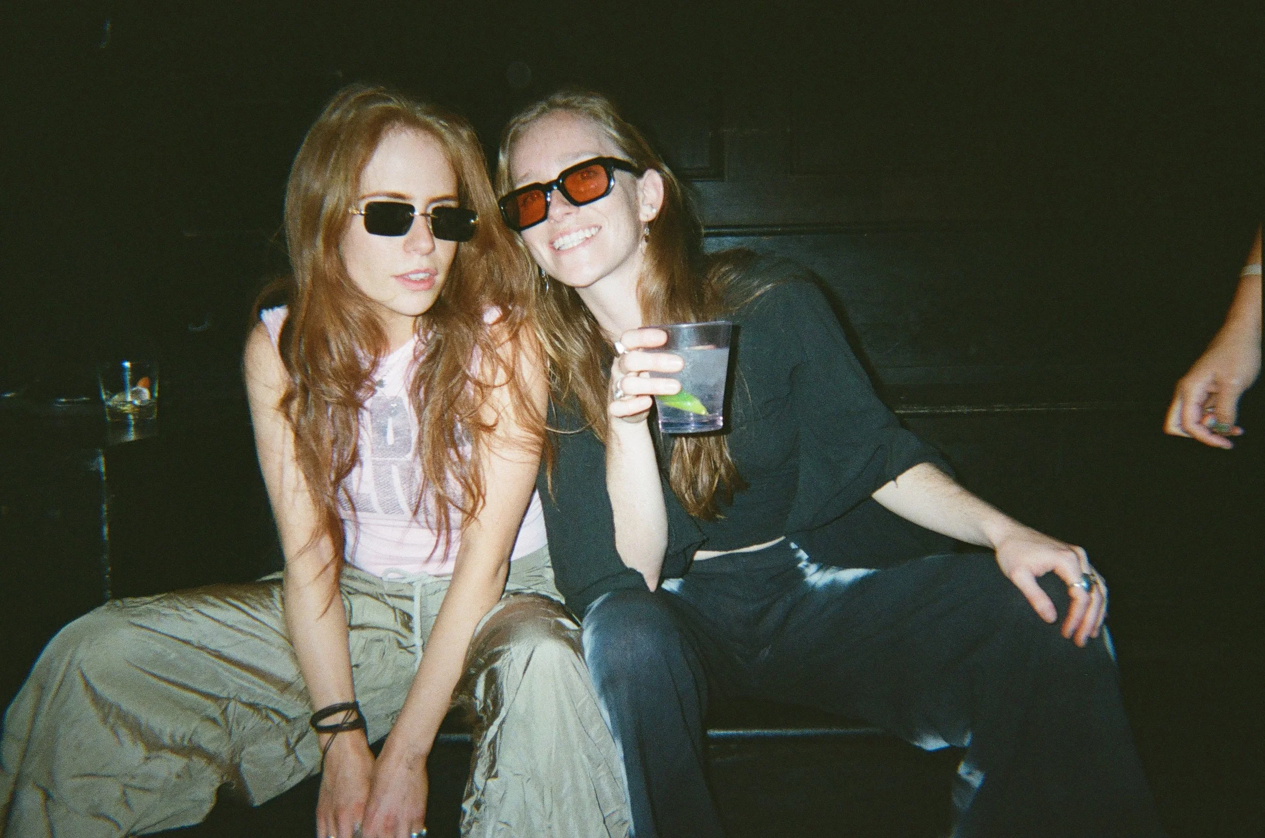 Two women sitting close together in a dimly lit setting, both wearing sunglasses. The woman on the left has red hair and is dressed in casual clothing, while the woman on the right has long hair and is smiling widely, holding a drink with lime.