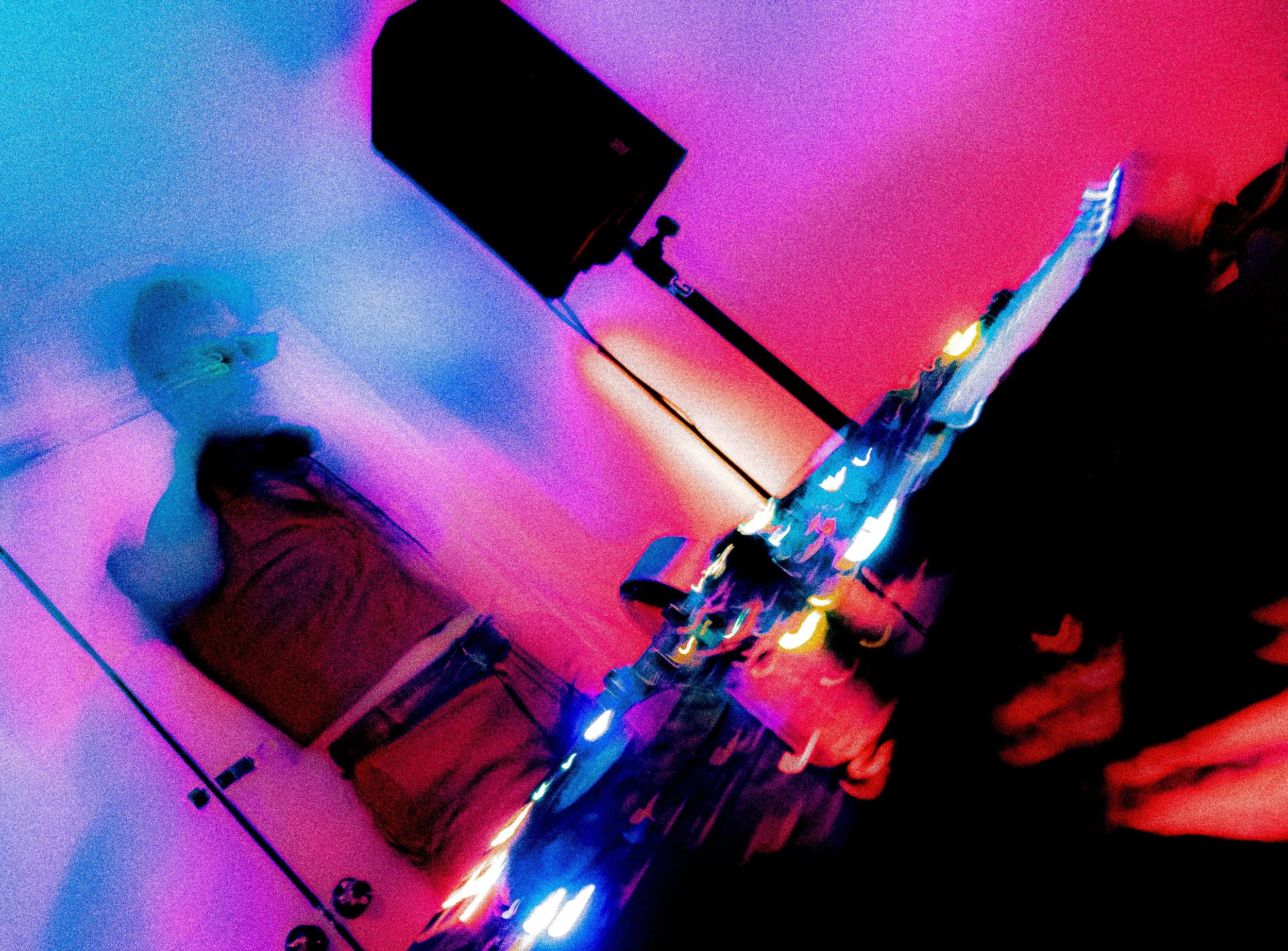 A person sitting at a desk in a room with colorful, neon purple, pink, and blue lighting. There is a computer monitor, a keyboard, a mouse, and decorative string lights on the desk. The person is wearing a red shirt and glasses, and appears to be wor