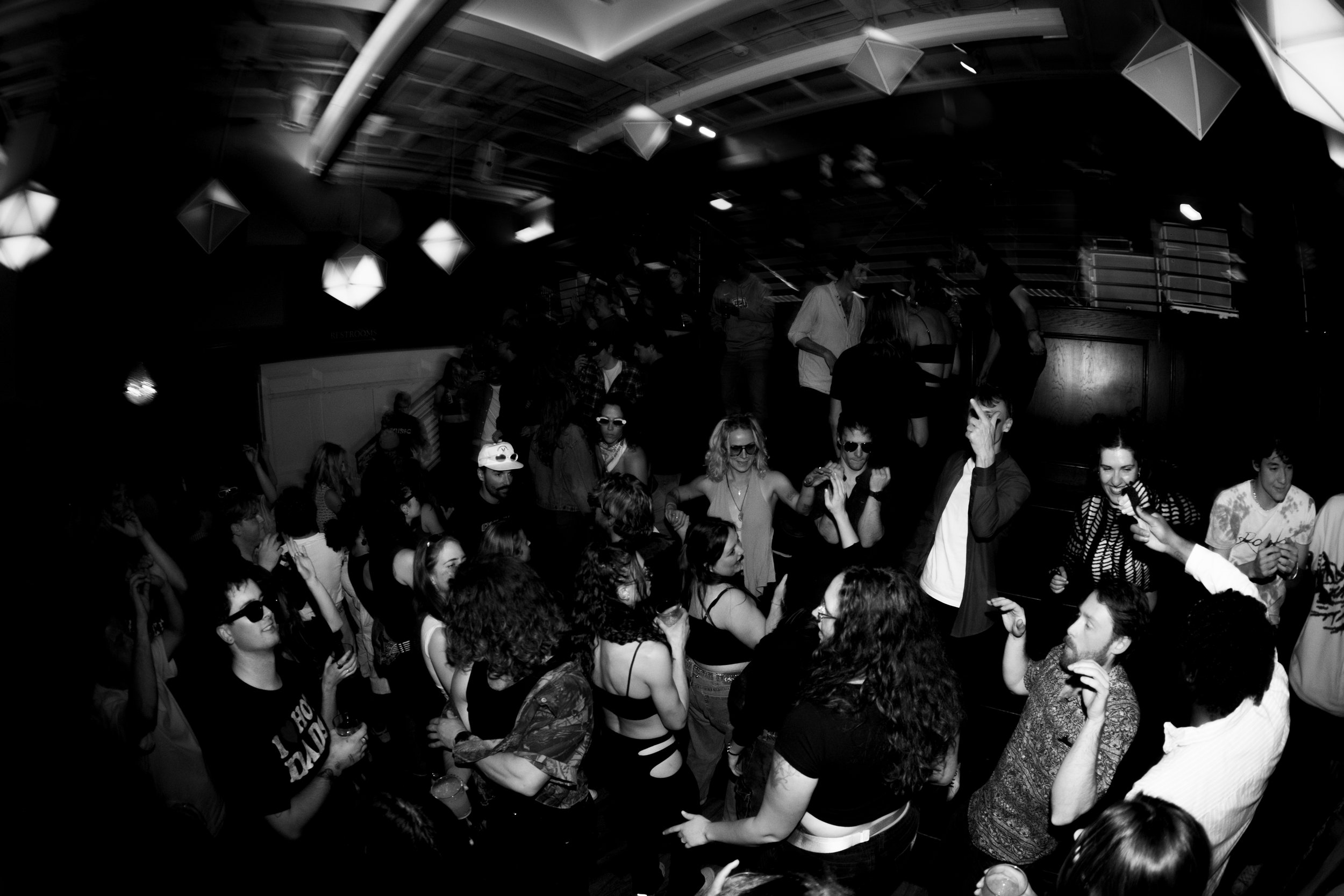 A black and white photo of a crowded dance floor with people dancing and socializing, illuminated by hanging geometric light fixtures.