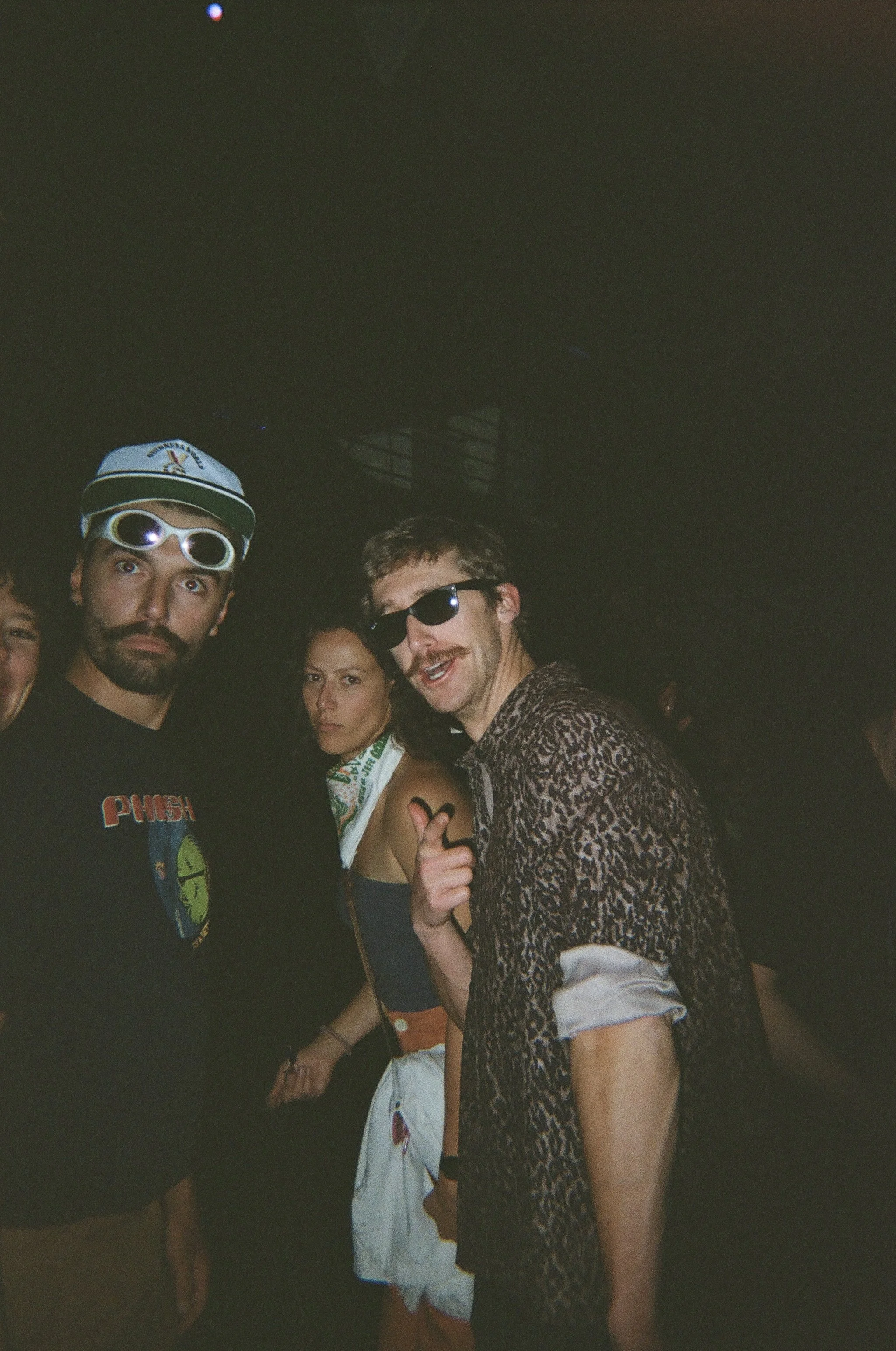 A group of four young people dressed in casual and festival attire, posing and smiling for a photo at night in a dark environment, with some wearing sunglasses and a baseball cap.