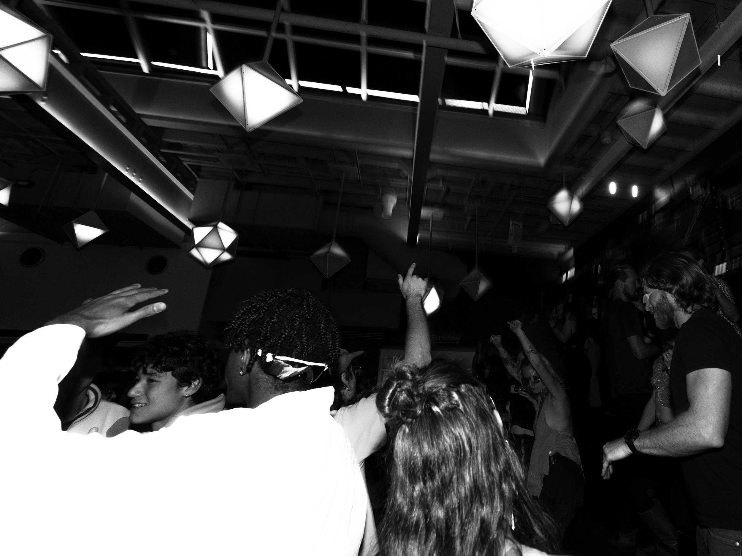 A black and white photo captures a crowd of people with diverse hairstyles and skin tones in a lively indoor setting, possibly a party or social event. Several individuals have their hands raised, and some are smiling or engaged in conversation. The 