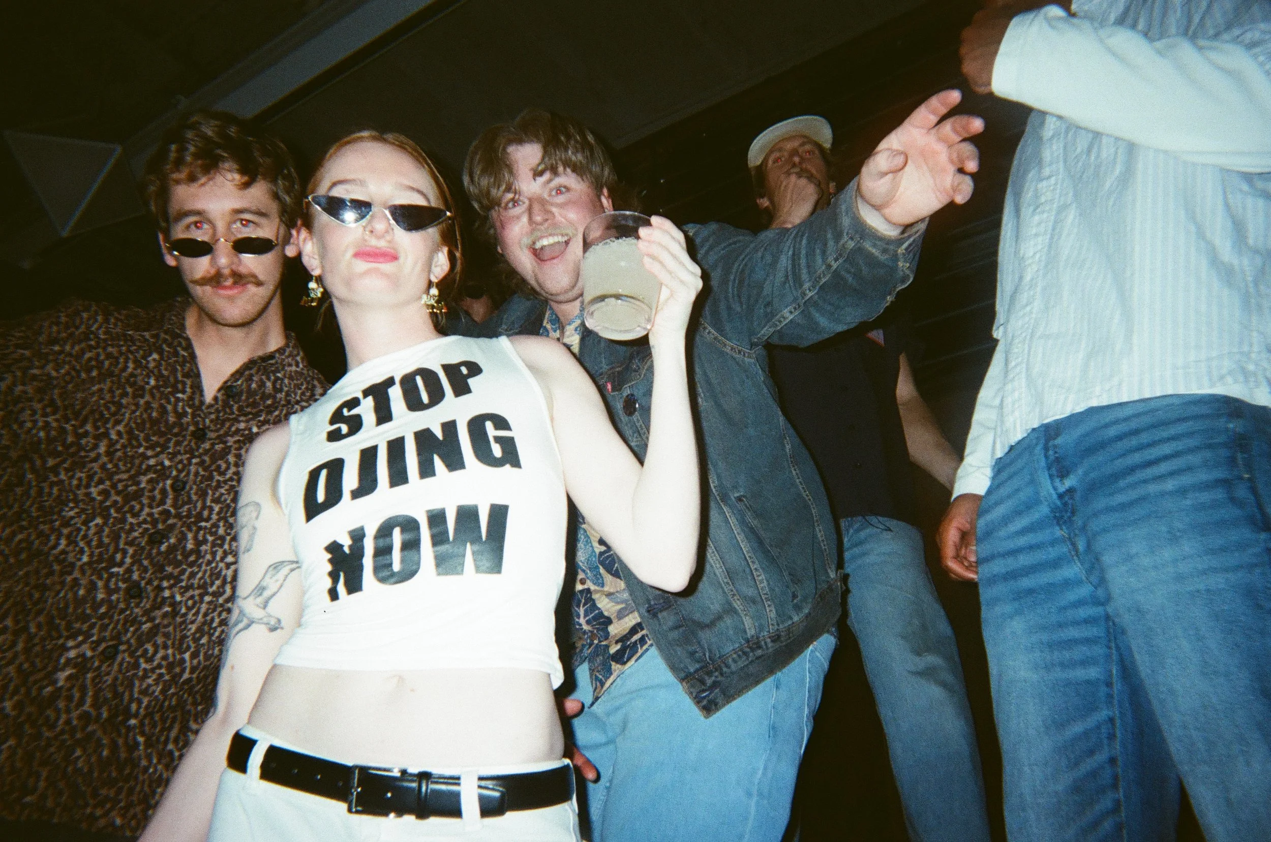 A group of four young adults at a party or club, with three people in the foreground posing and smiling at the camera, and one person in the background covering their mouth.