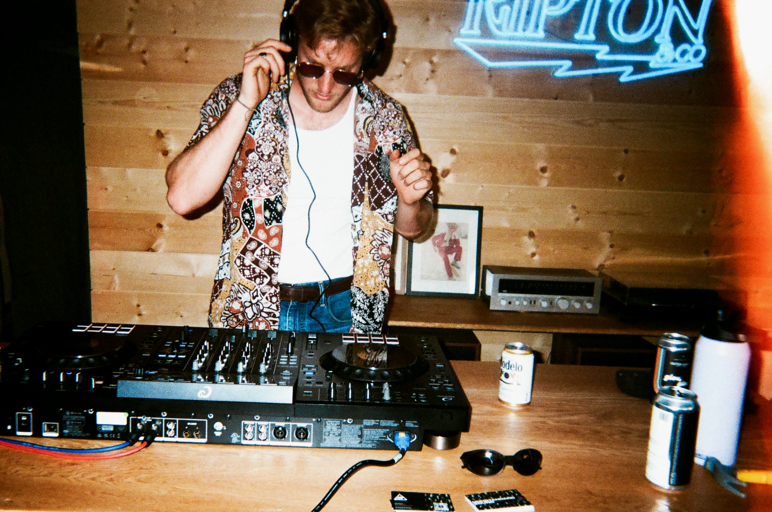 A man with sunglasses and a patterned shirt is DJing, adjusting headphones, standing behind a DJ controller in a room with wood-paneled walls. There are framed pictures and audio equipment on the wall behind him, with neon signage partially visible.