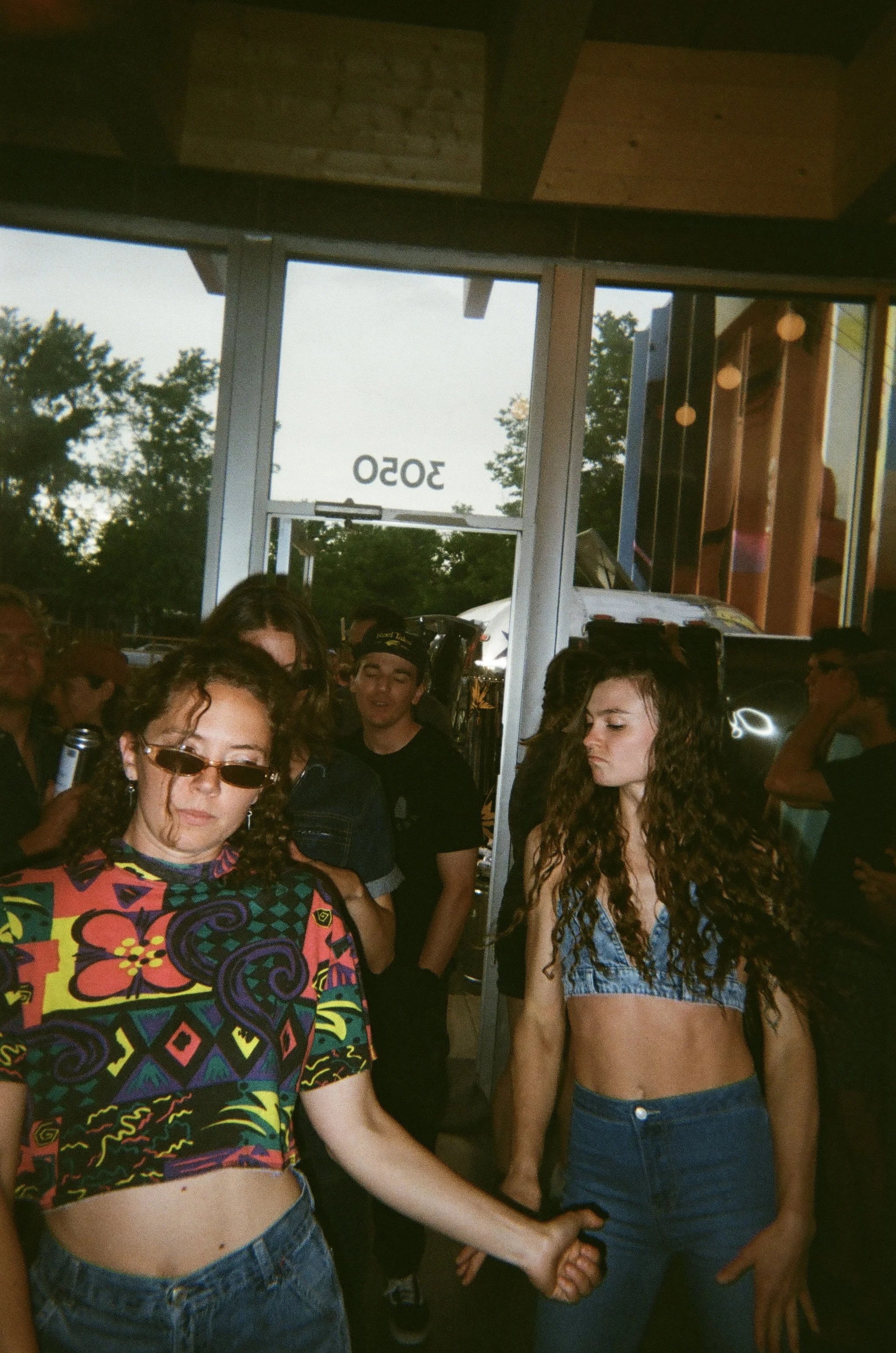 Group of young women with curly hair in casual outfits, holding hands, inside a store or cafe with glass doors and people in the background.