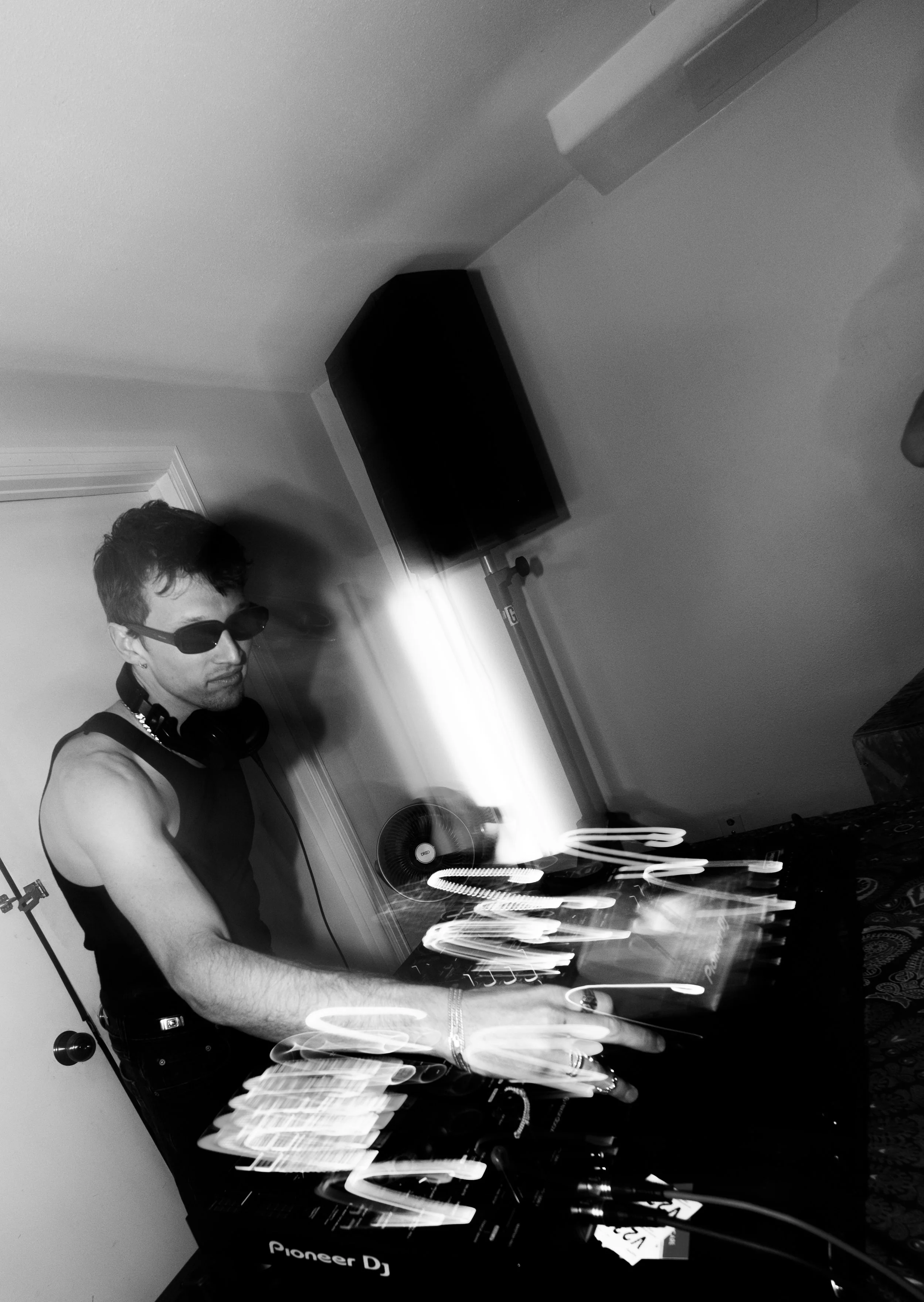 A person wearing sunglasses and headphones is DJing at a setup with turntables and a mixer, in a room with a wall-mounted speaker and a fan.