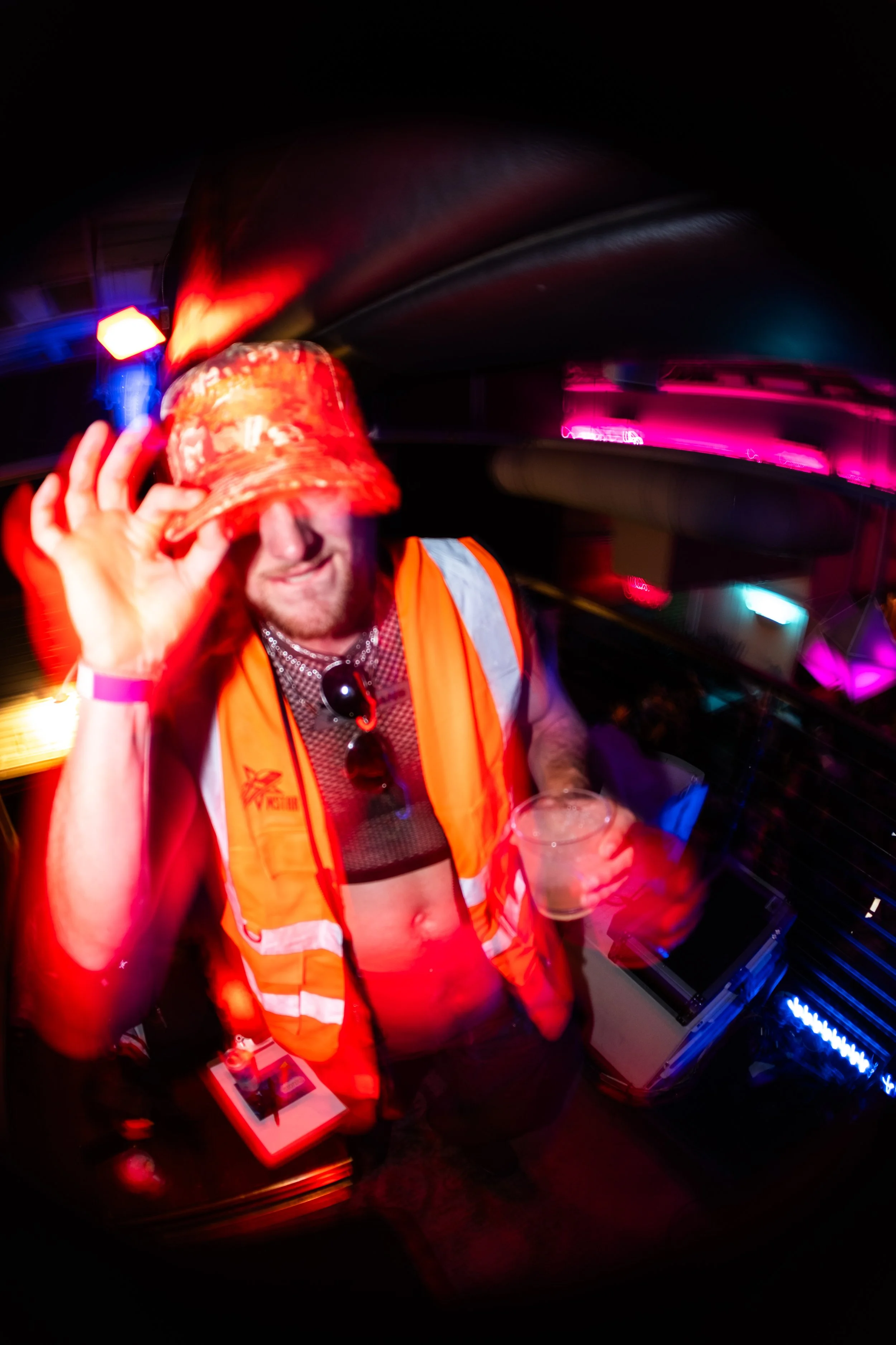 Person wearing a bright orange safety vest and red bucket hat, holding a plastic cup while touching the brim of the hat, at a nighttime event with colorful lights and electronic equipment.