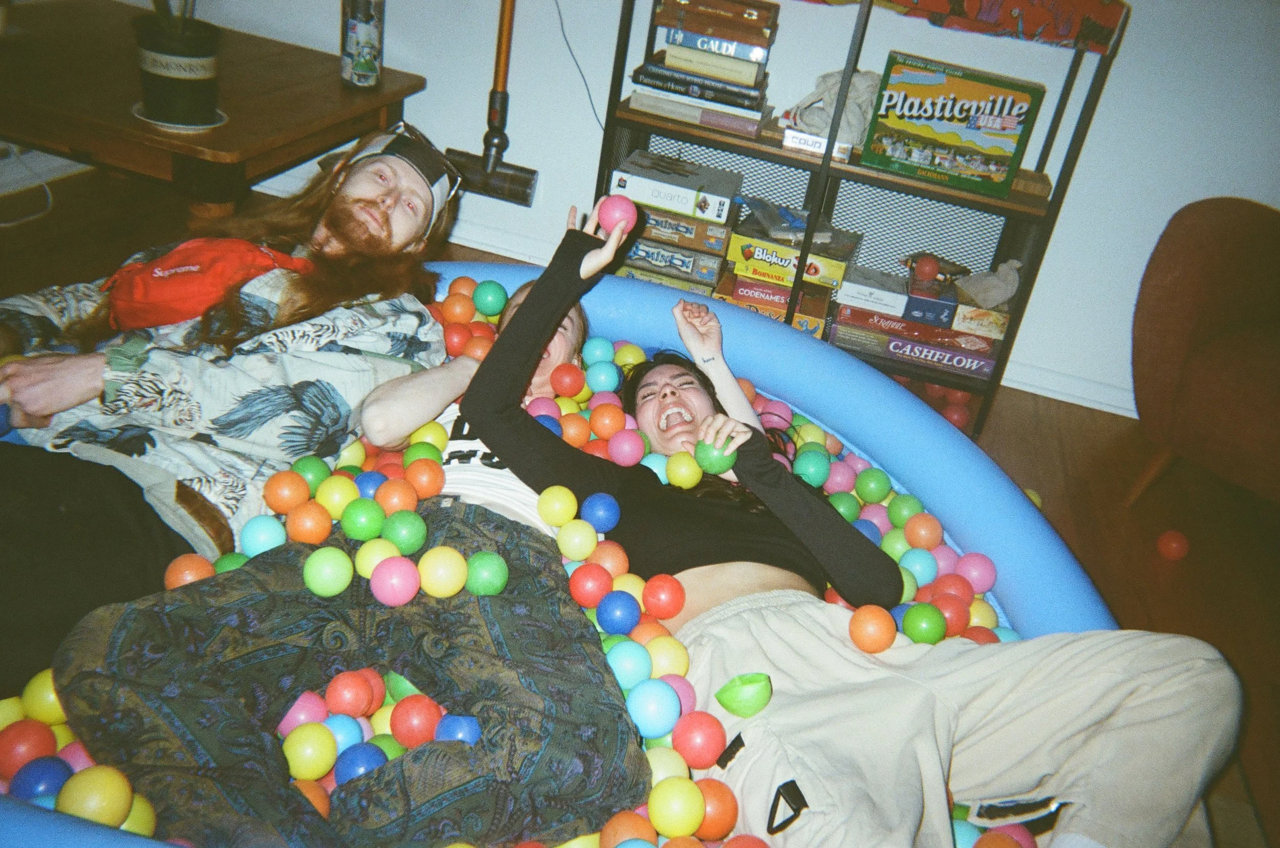 Three people lying in a ball pit filled with multicolored plastic balls, smiling and having fun indoors near a bookshelf filled with board games and books.
