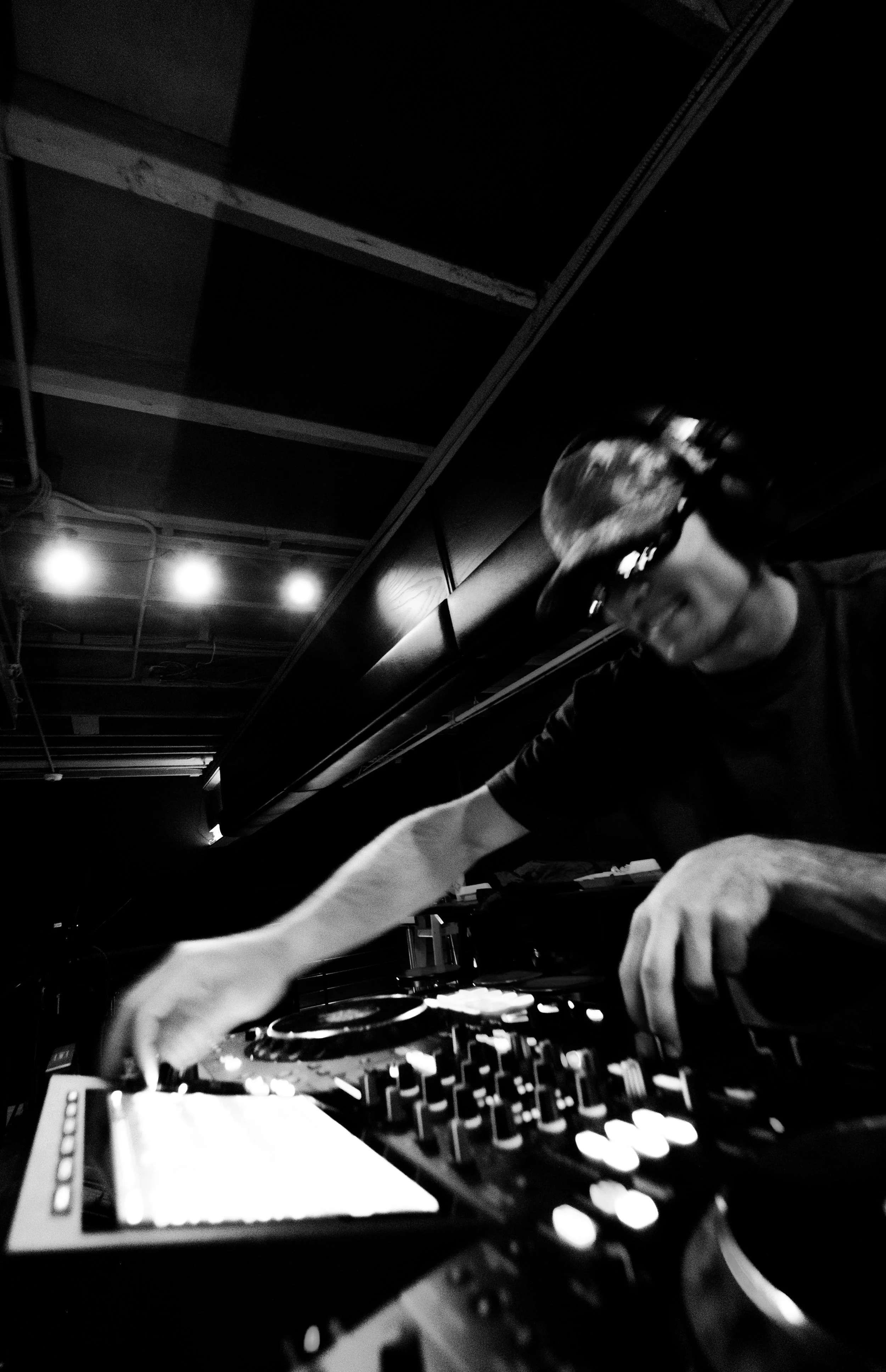 A DJ wearing headphones, leaning over a mixing console, in a dark nightclub setting with ceiling lights.