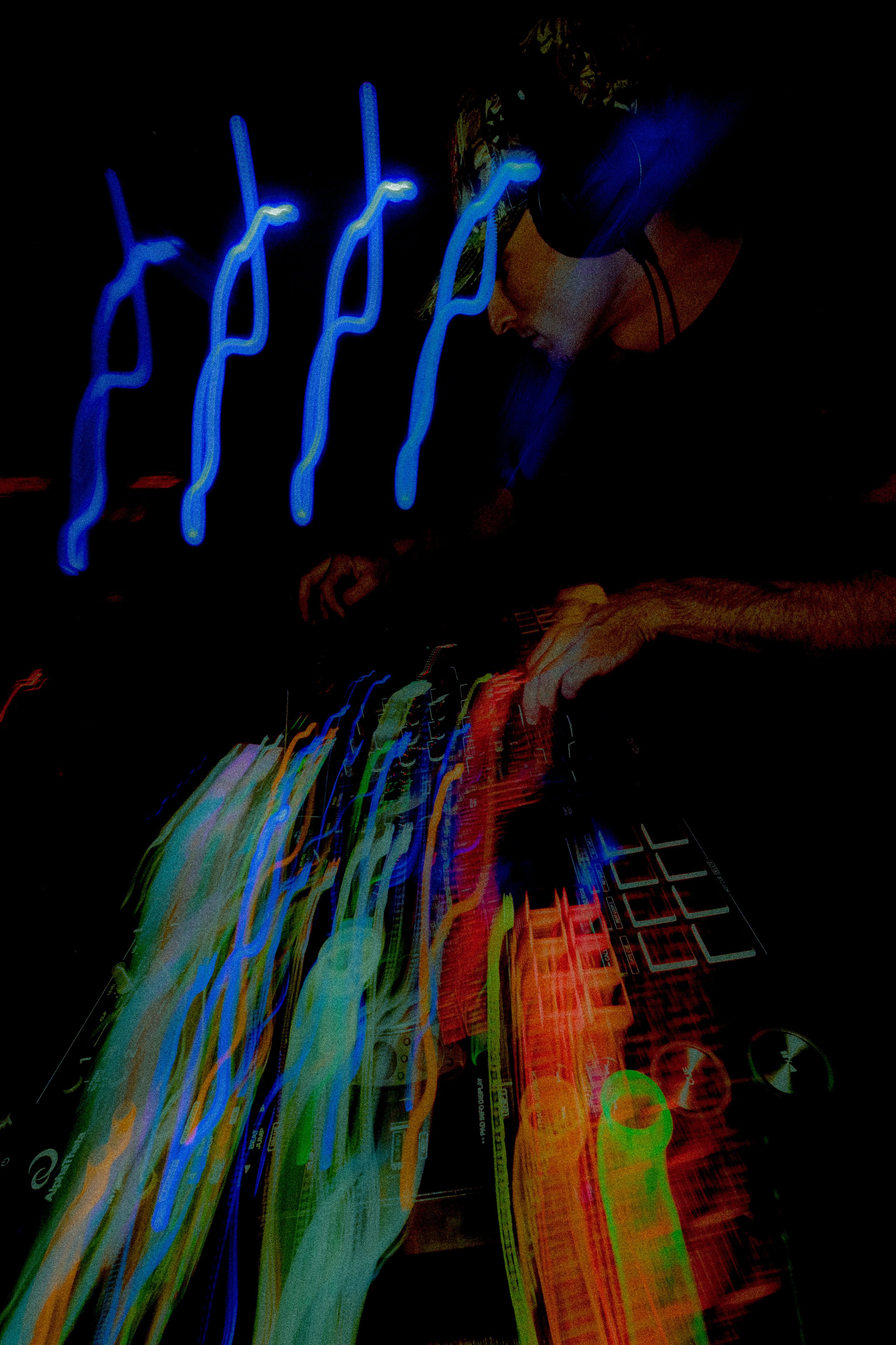 A person wearing headphones appears to be DJing or working with music equipment. The image has long exposure light trails in various colors, creating a vibrant, abstract visual effect.