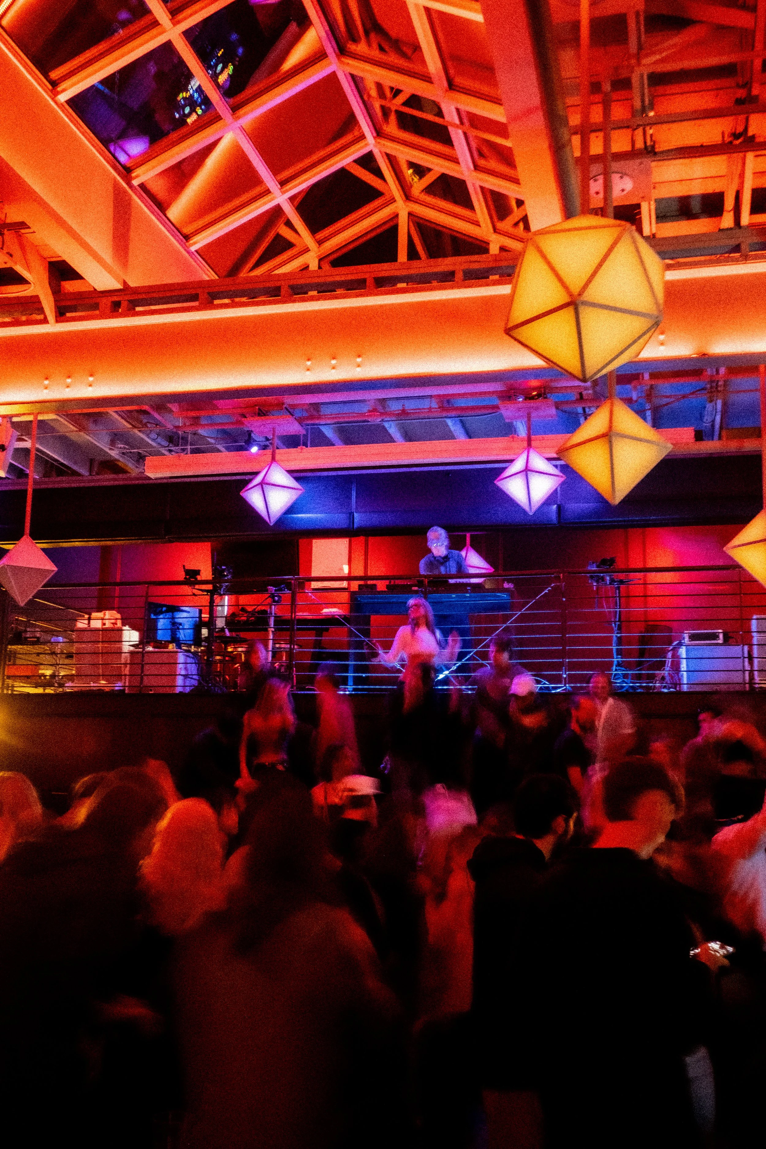 A crowded dance floor with people dancing under colorful hanging lamps in a venue with a modern ceiling design and a DJ playing music.