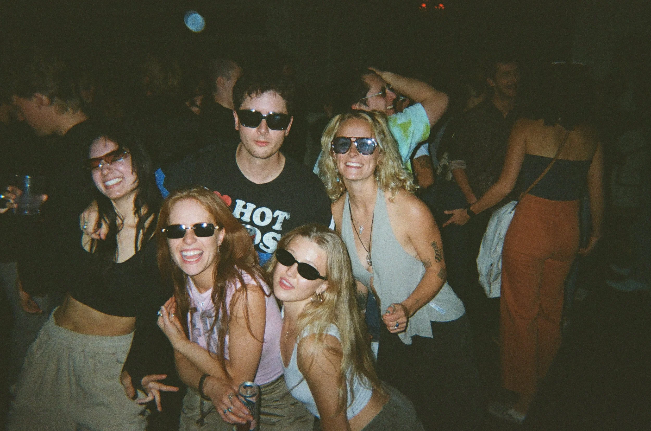 Group of young people dancing and having fun at a party or nightclub, wearing sunglasses and casual summer clothes.