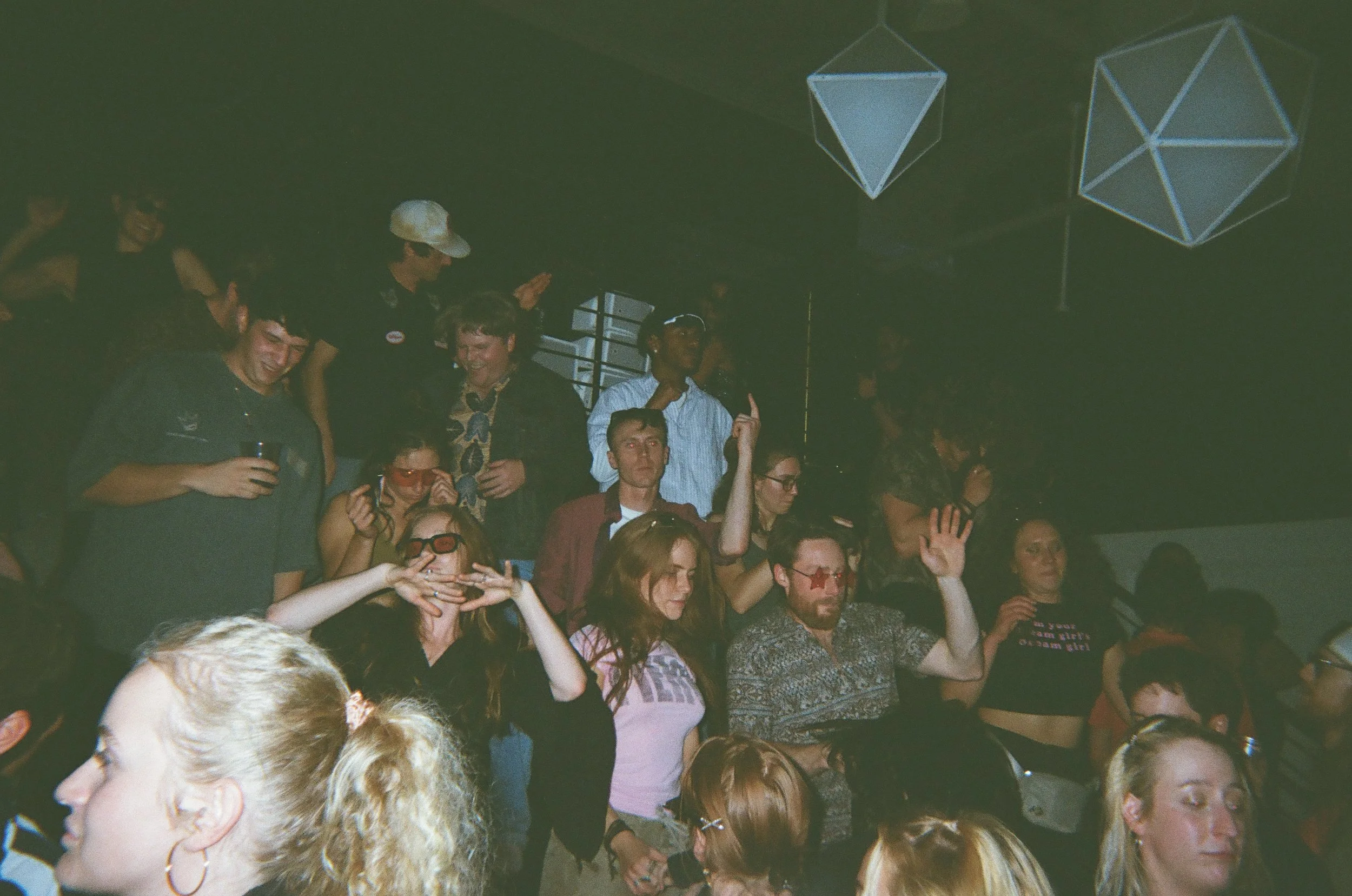 Crowd of people at a party or concert with some raising their hands and others holding drinks, with geometric hanging decorations in the background.