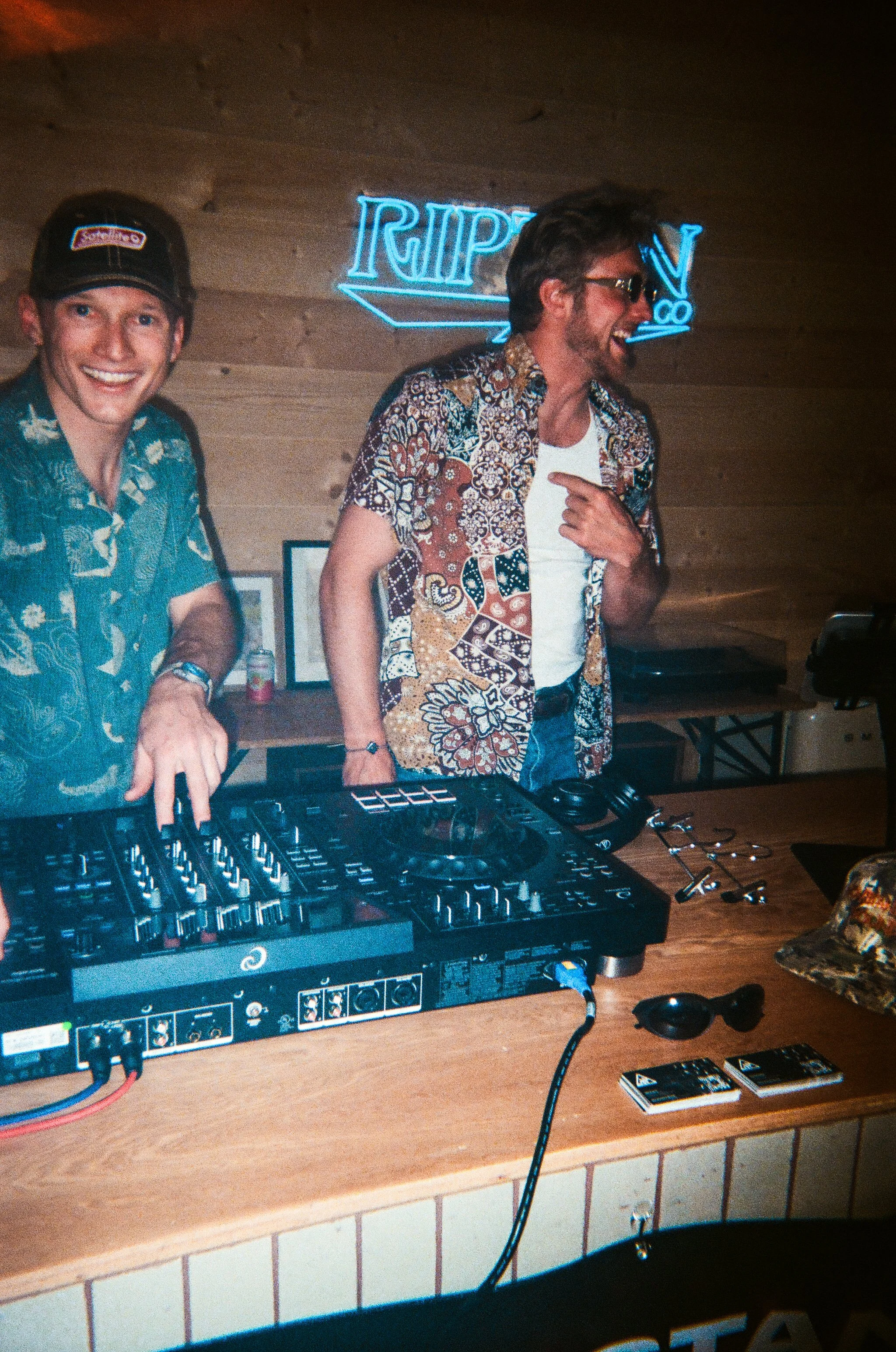 Two men enjoying music at a bar or club with a DJ setup on a wooden table in front of them. One man is smiling and wearing a patterned shirt and a cap, while the other man is laughing, wearing glasses and a colorful shirt. 