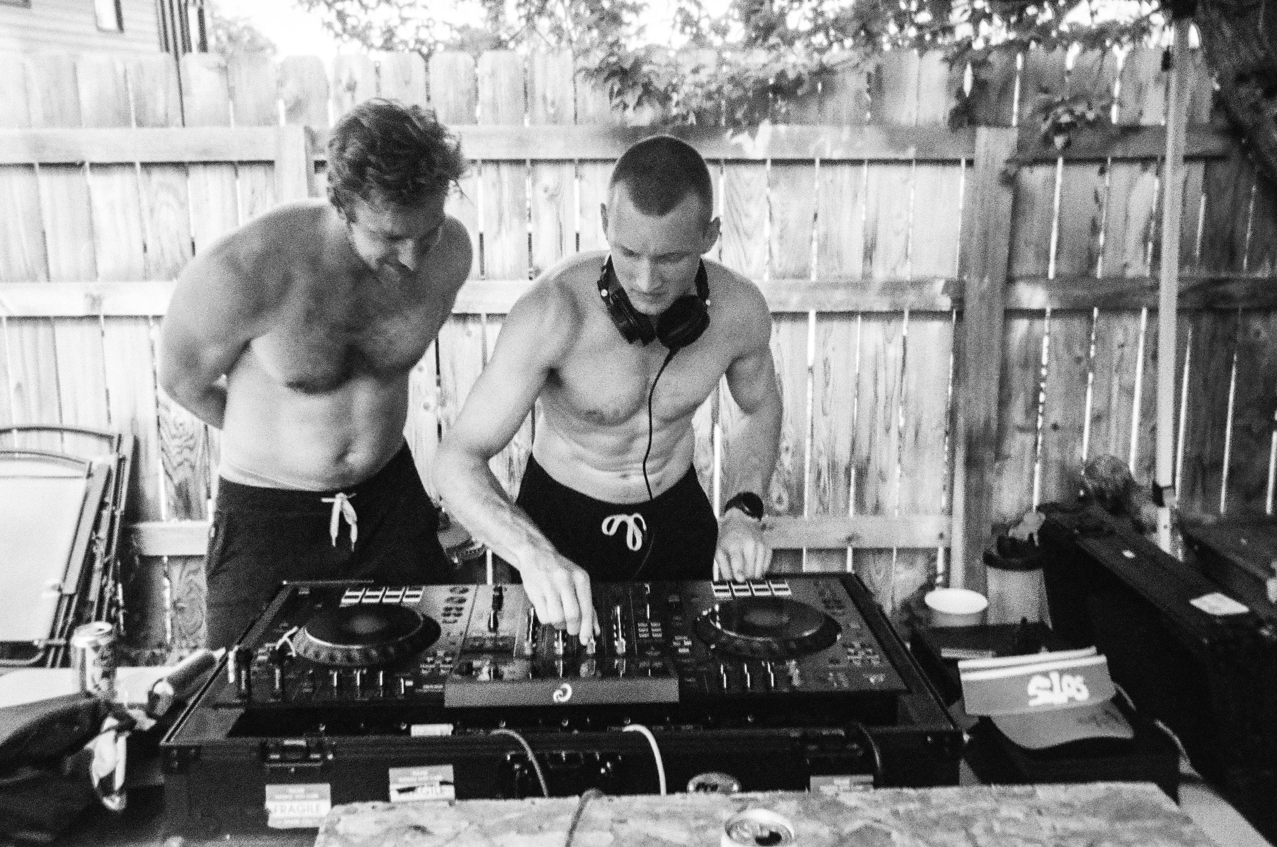 Two shirtless men with short hair and tattoos are DJing outdoors behind a DJ turntable setup. One man is wearing headphones around his neck, and they are focused on the music equipment. The scene is in a backyard with a wooden fence and some outdoor 