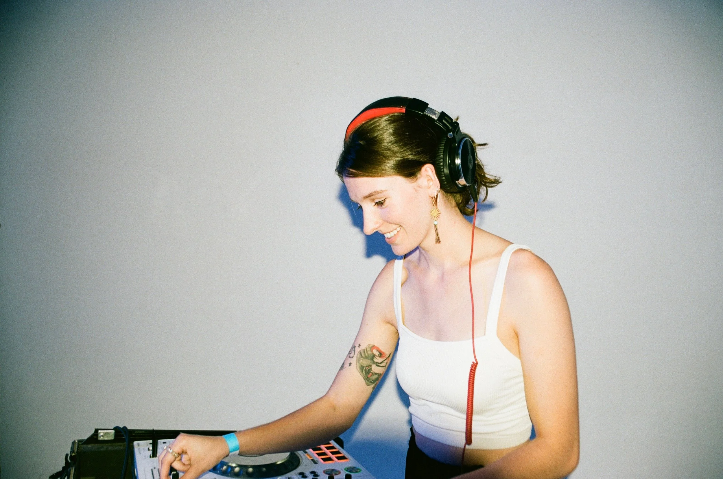 A woman with short dark hair, wearing headphones and a white tank top, smiling as she DJs with a mixer and electronic equipment in front of her.