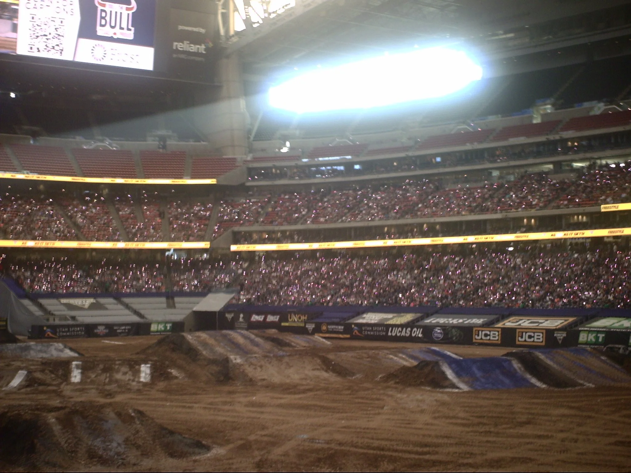 View of an indoor stadium with a dirt track in the foreground and a crowd of spectators in the stands. Bright stadium lights shine from above, and various city and sponsor banners are displayed around the upper levels.