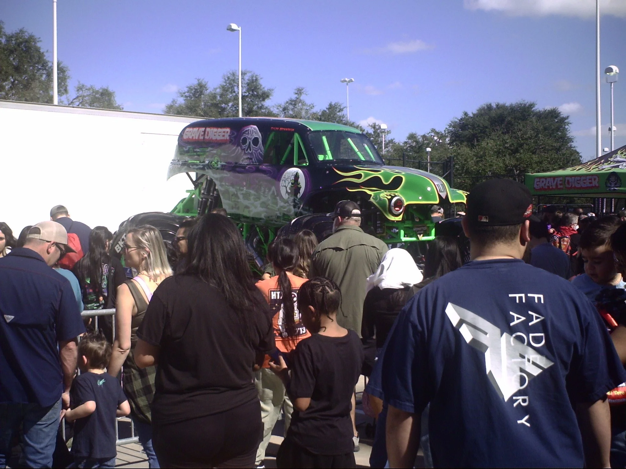 A crowd of people gathered outdoors watching a monster truck with green and black flames and a skull design on its side. The sky is blue with some clouds, and there are tall lampposts in the background.