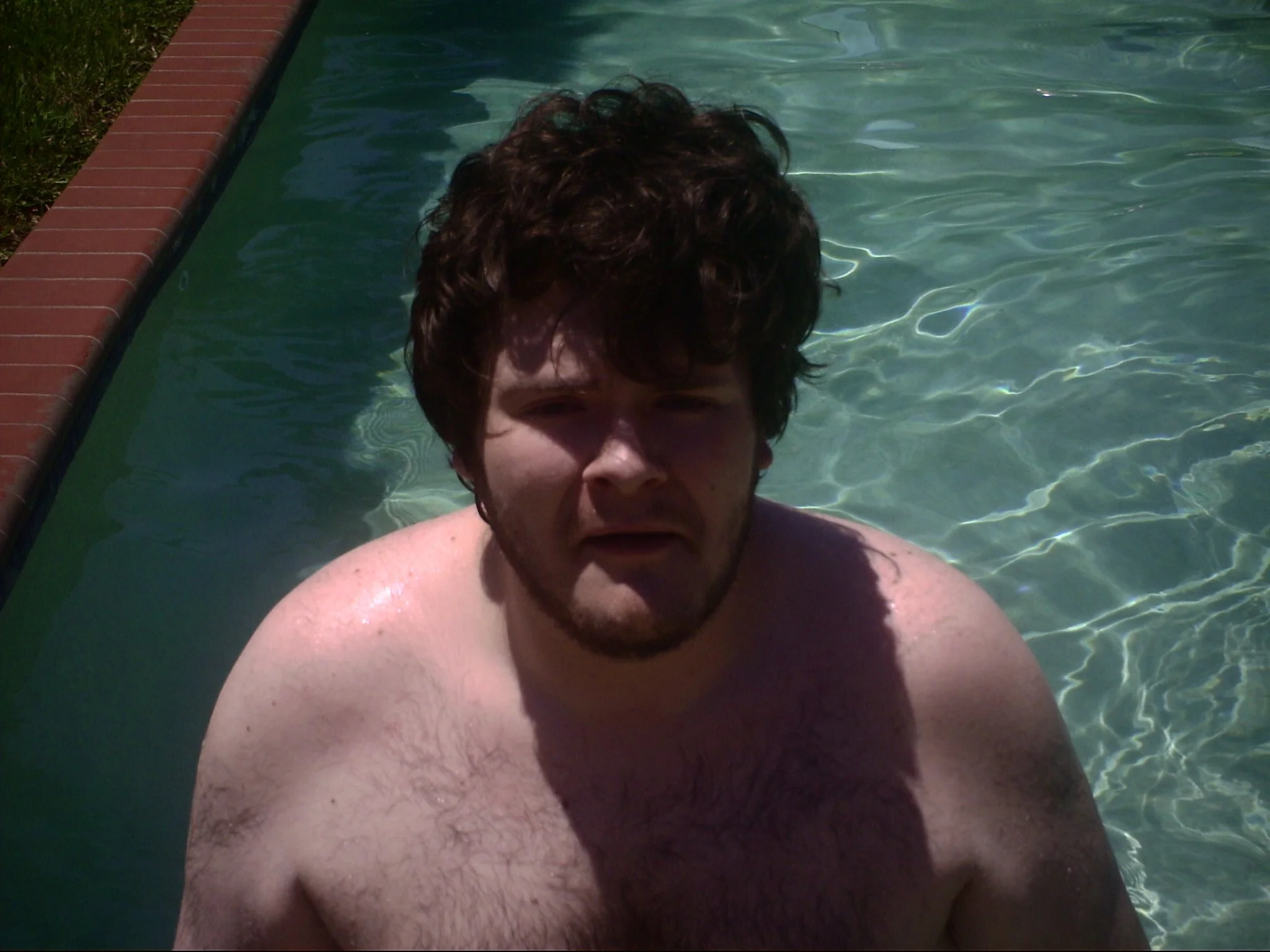 A person with curly hair and facial hair taking a selfie in a swimming pool, with water and a brick edge visible.