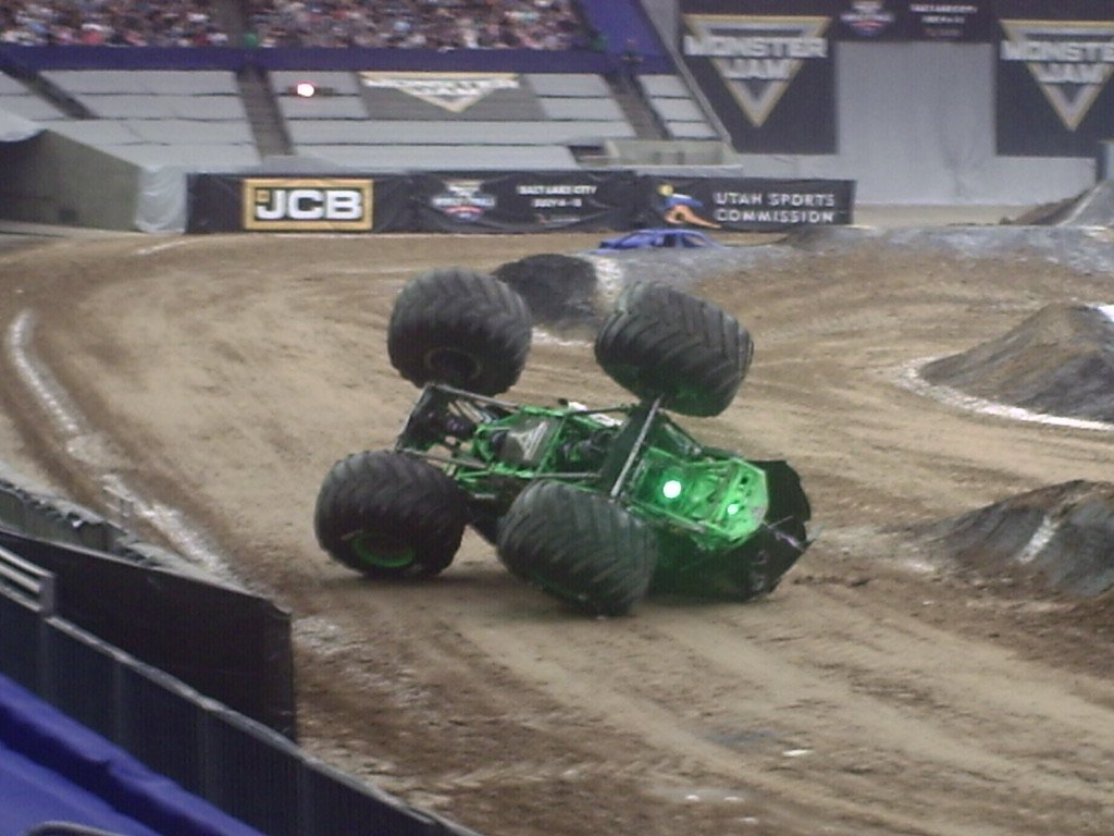 A green monster truck is flipping upside down on a dirt race track at an indoor stadium.