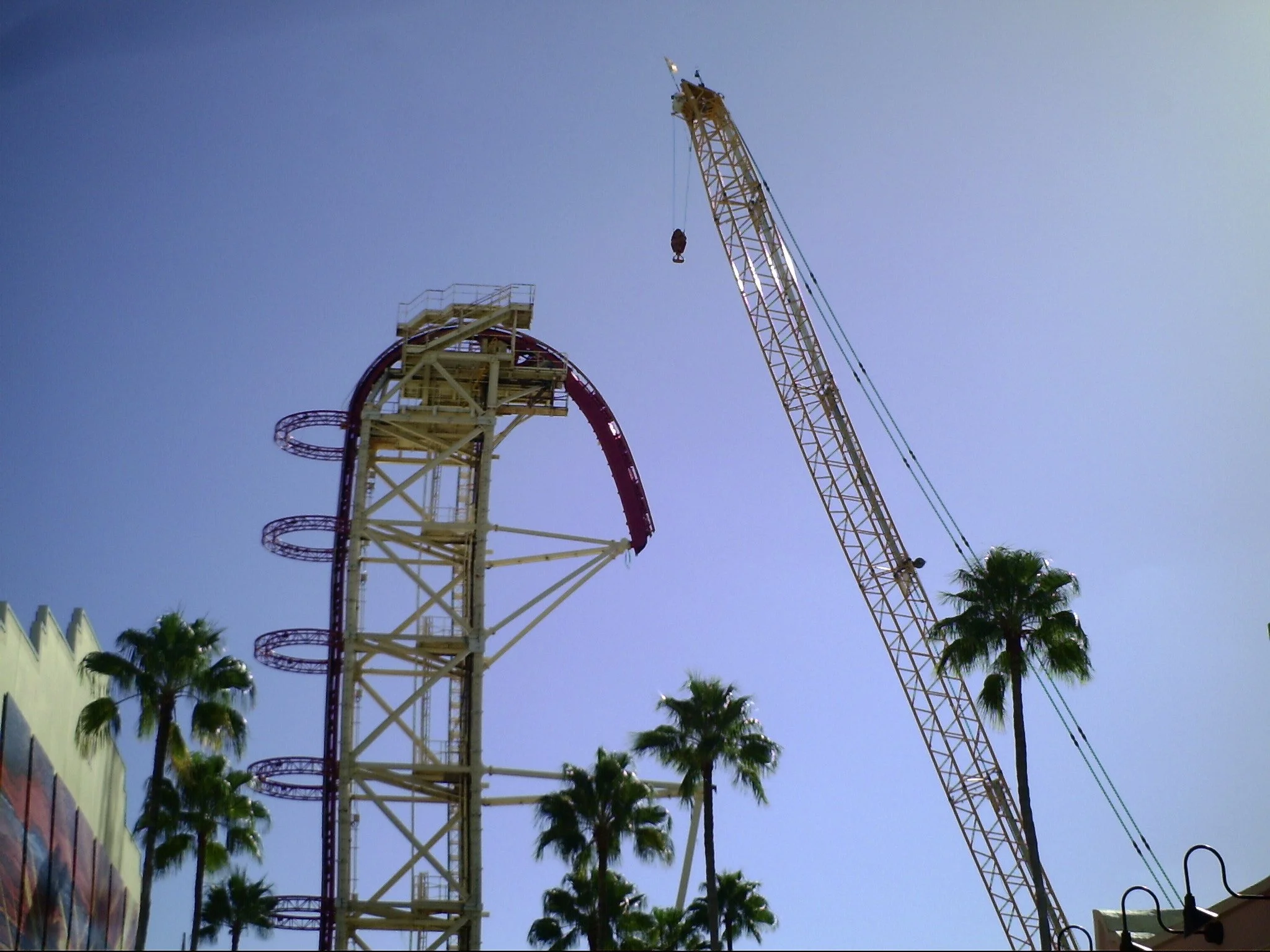 A roller coaster with yellow supports and red tracks, a crane, tall palm trees, and a building with a white wall and colorful panels, under a clear blue sky.