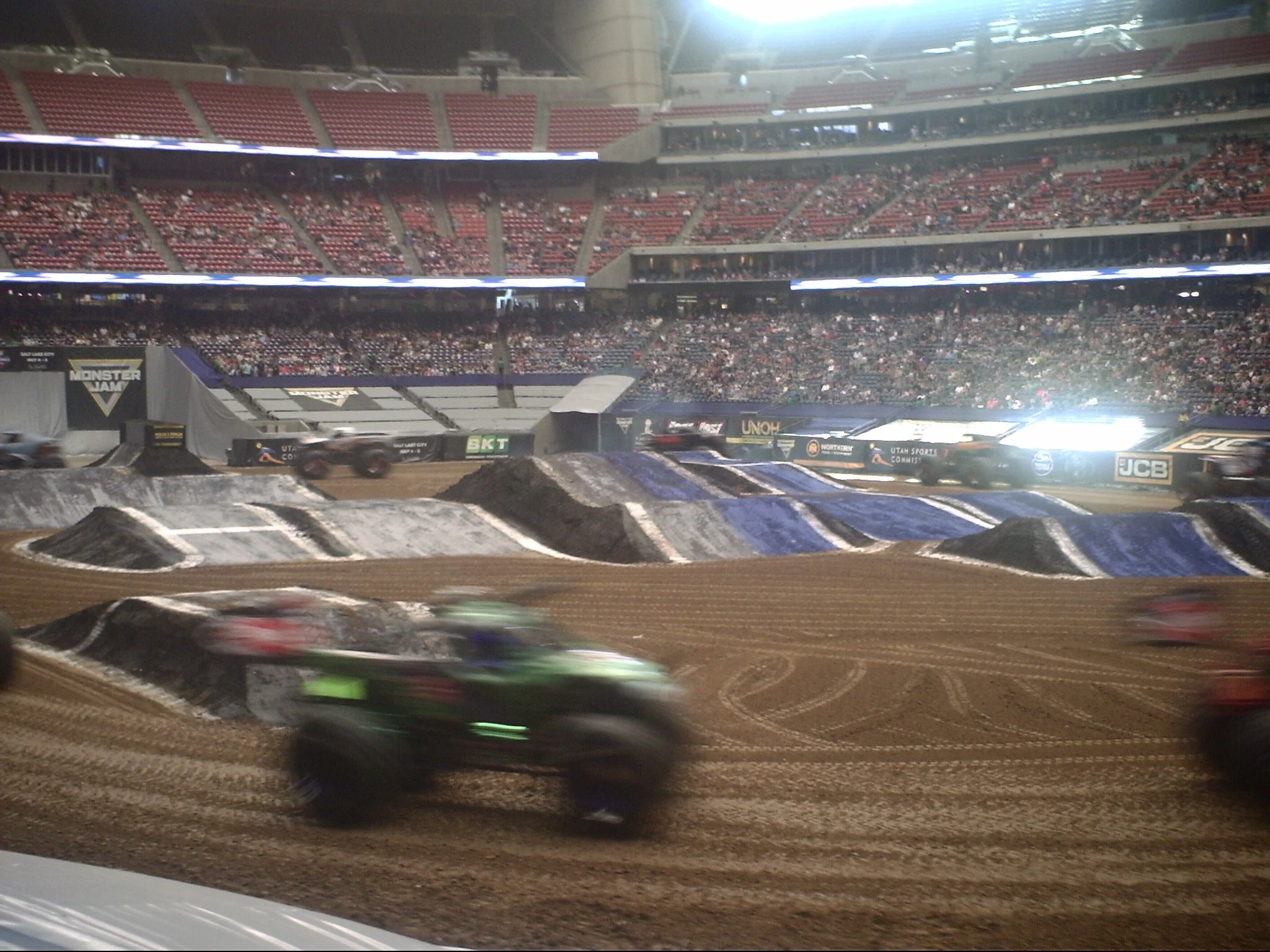 Racing cars speed around a dirt track inside a large stadium with many spectators and bright lights overhead.