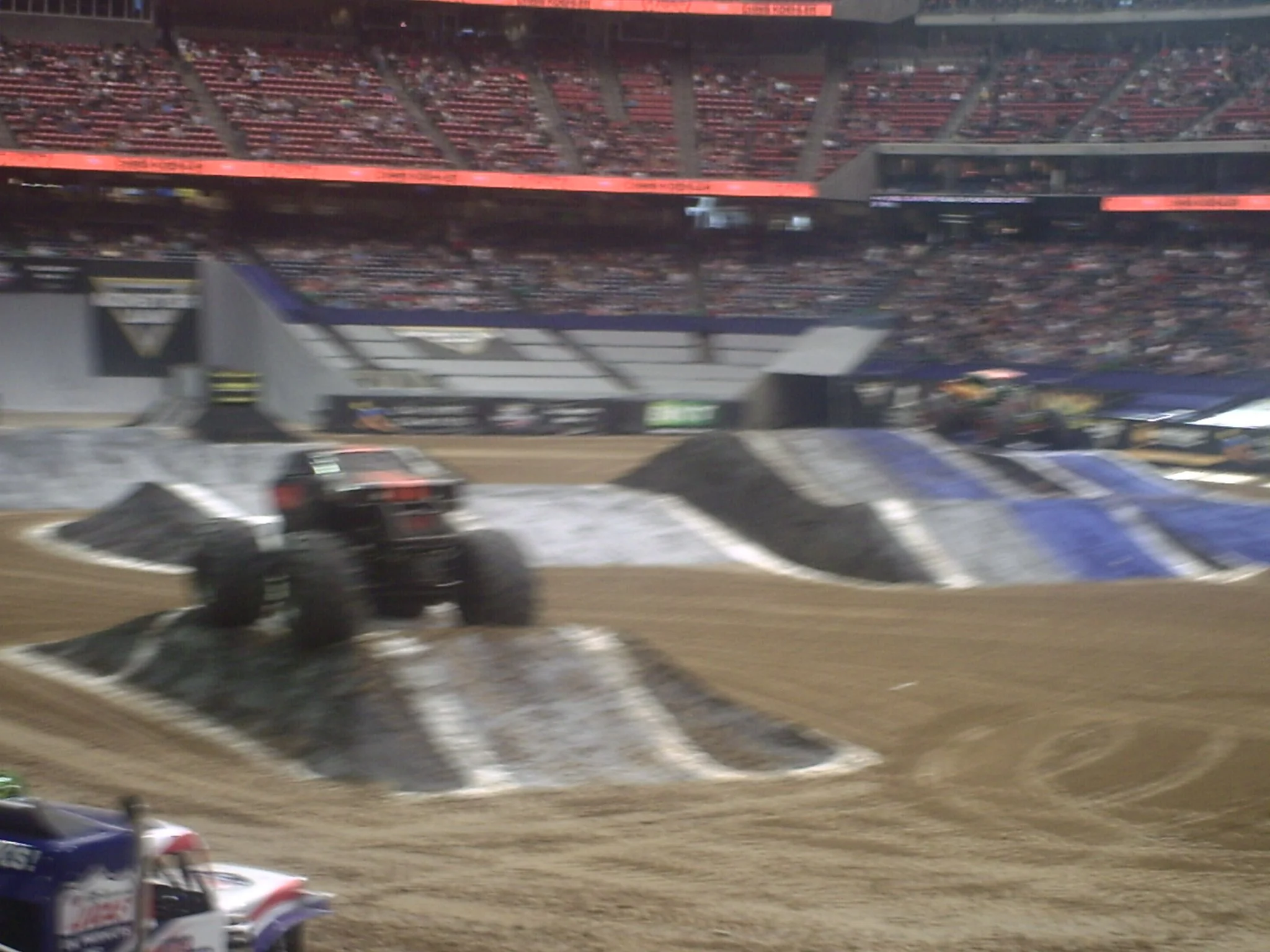 The image shows a stadium with a dirt racetrack and several race cars. Spectators are seated in the stands, and the scene appears to be during a nighttime event.