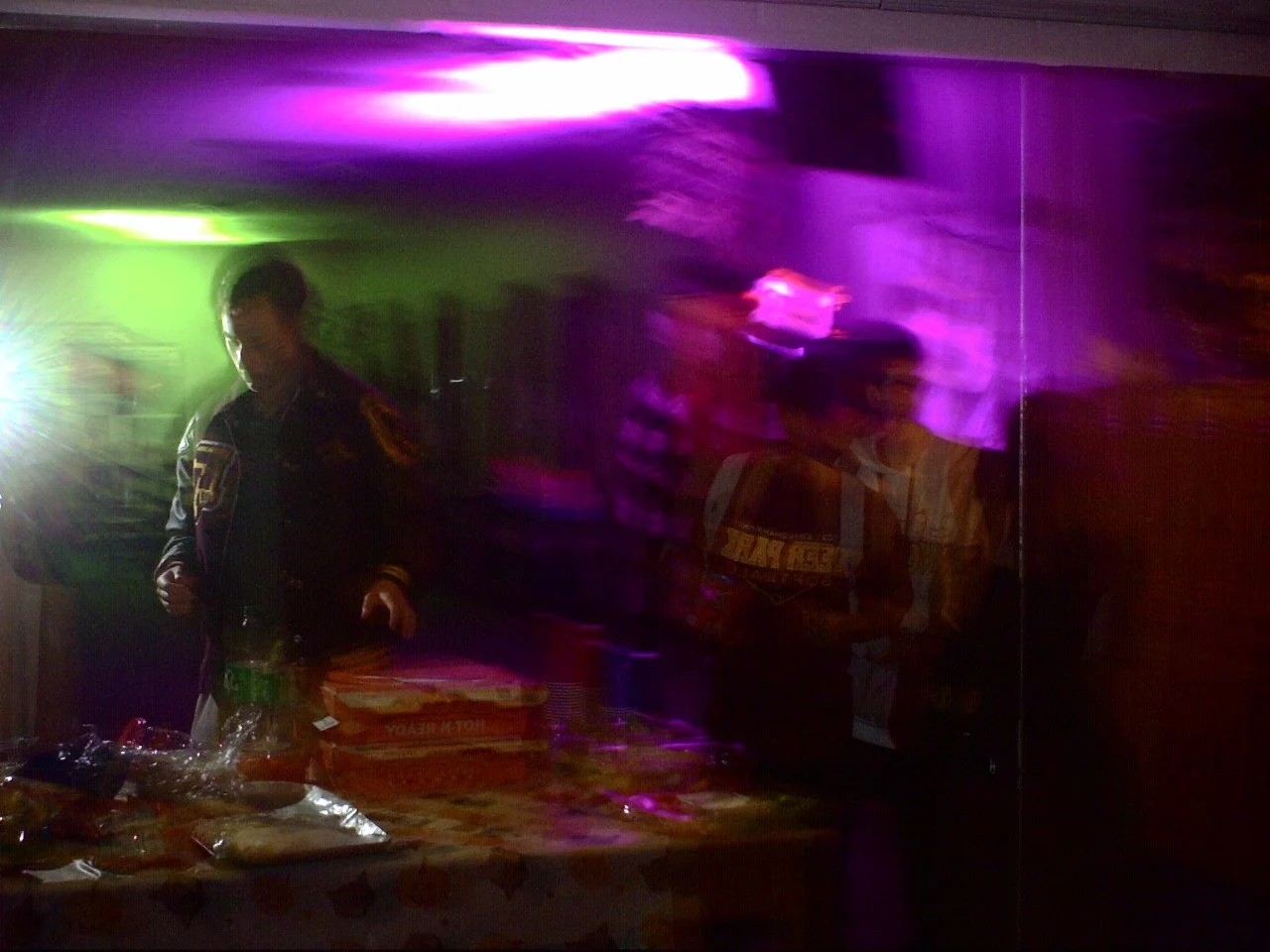 Dimly lit room with colorful purple and green lights, man in dark jacket, and a table with pizza boxes and snacks.