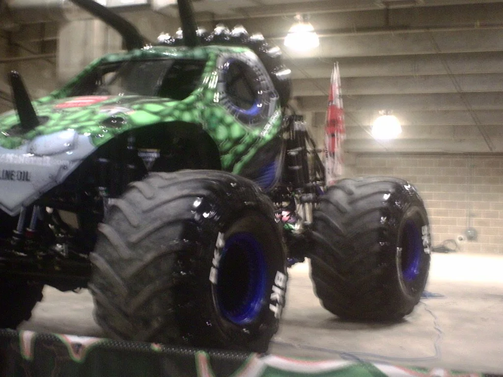 A monster truck with large tires, painted green and black, in an indoor garage or warehouse with overhead lighting and brick walls.