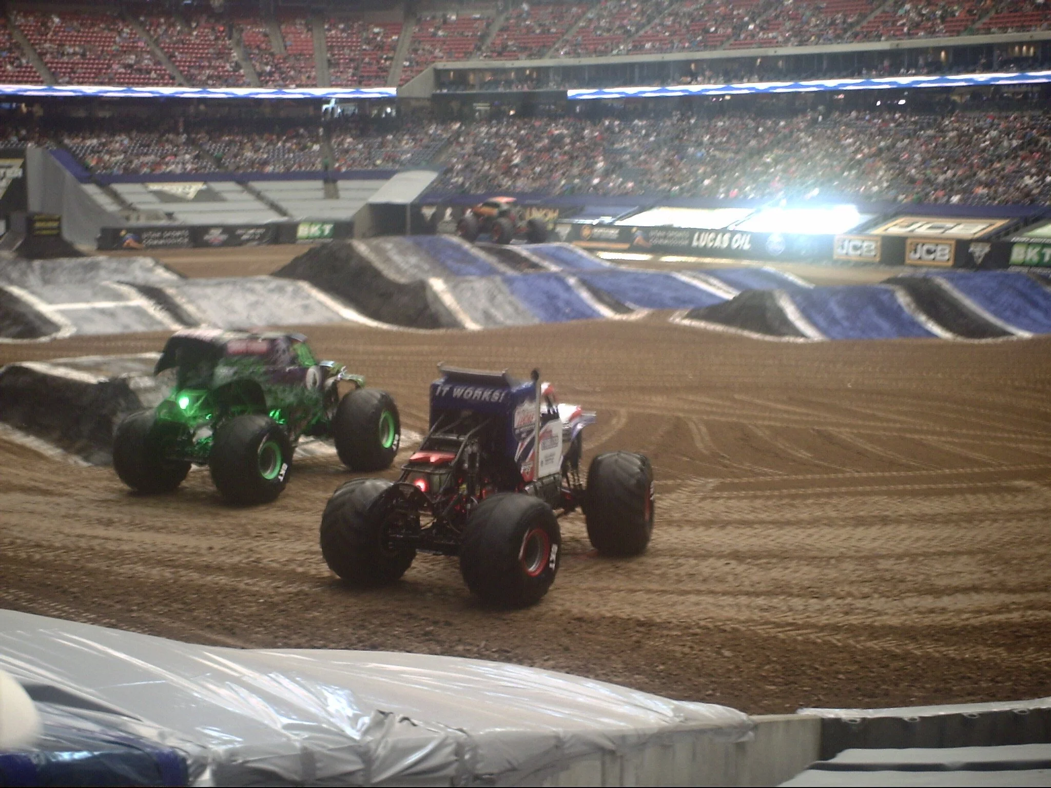 Monster trucks on a dirt track with a packed stadium audience watching.