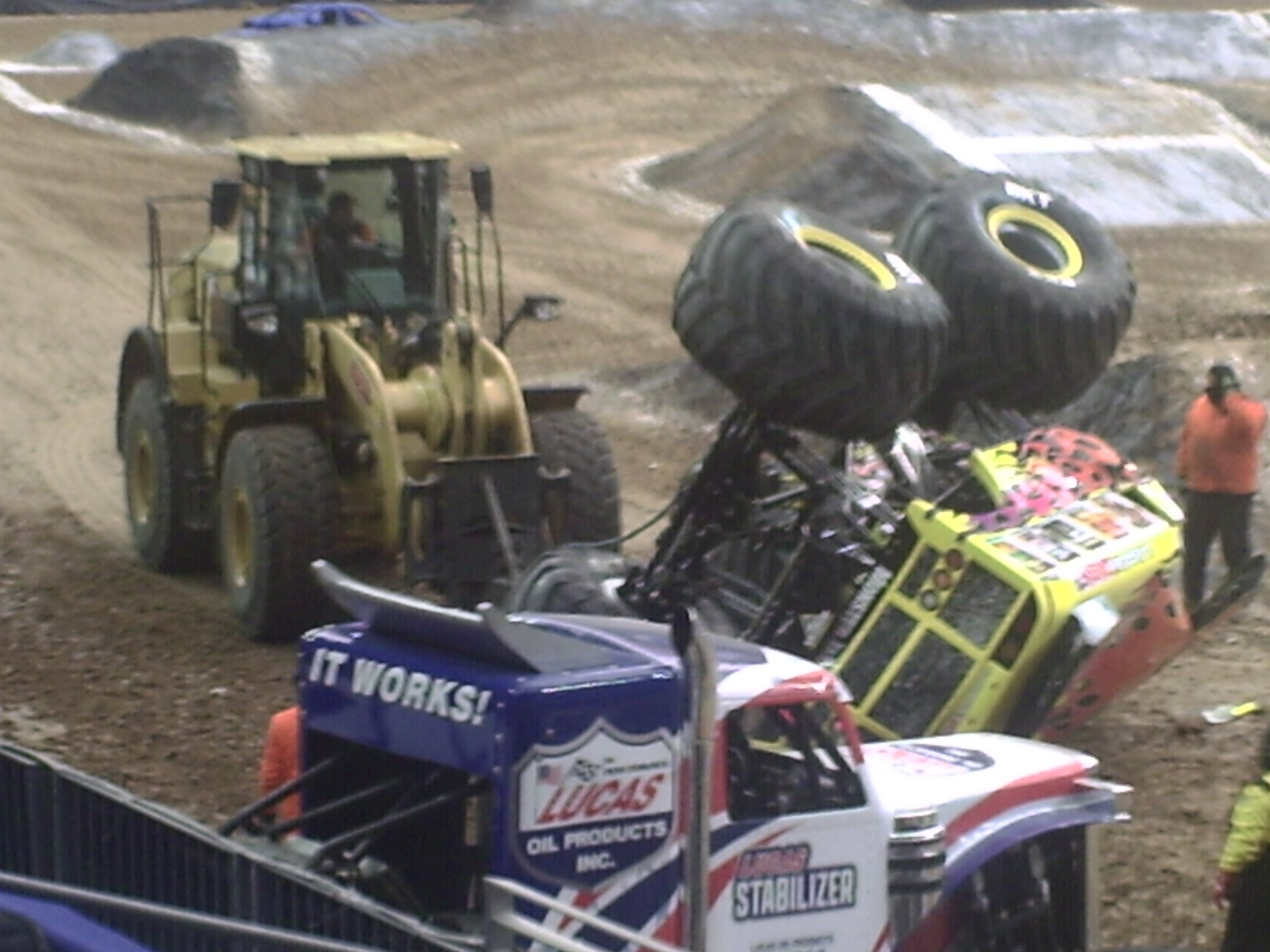 A dirt racetrack with racing vehicles, including a bright yellow and blue race car and a yellow bulldozer with a driver inside. There are multiple tires, some stacked, on the track and in the surrounding area.