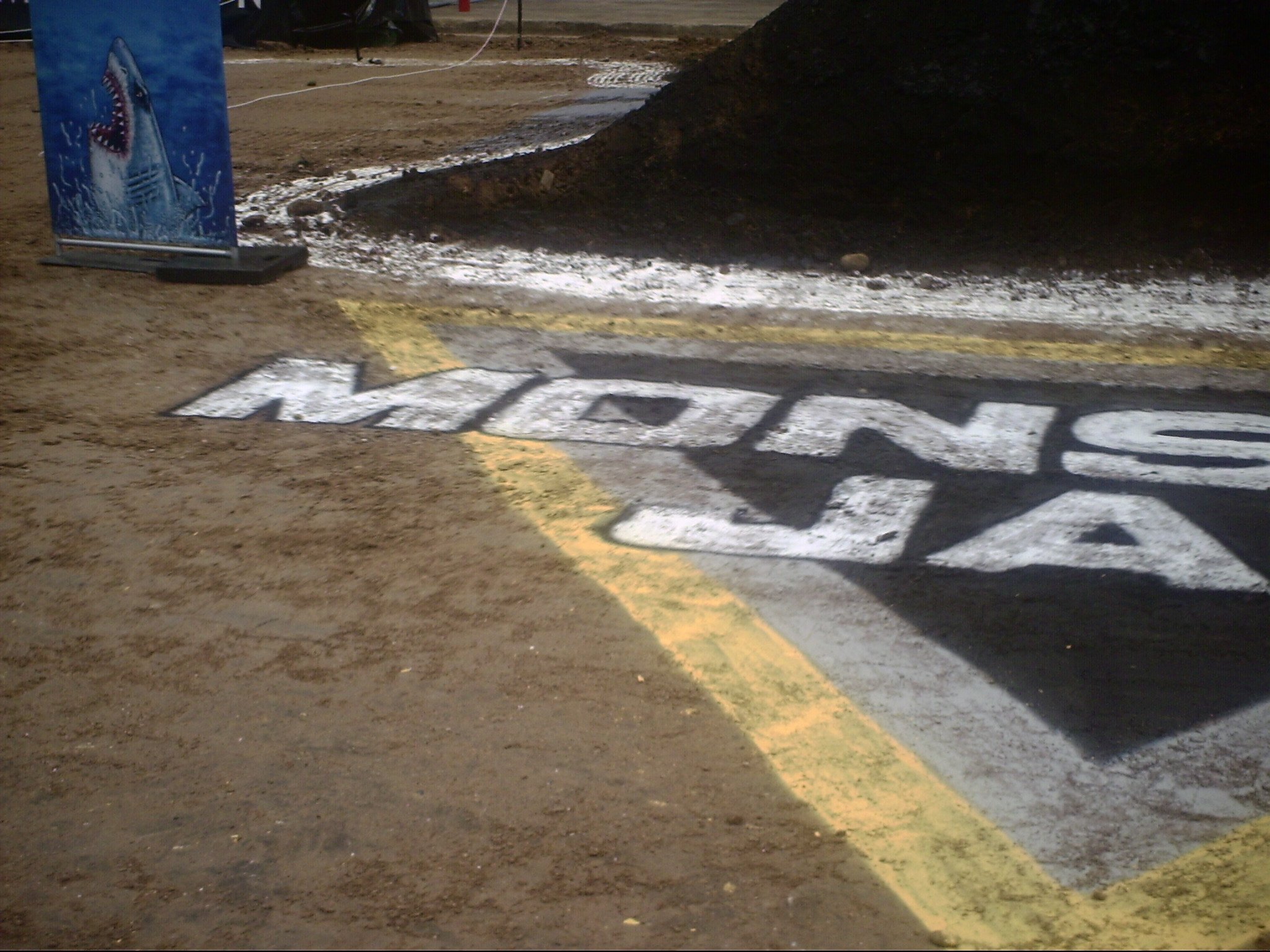 A construction area with a flood warning sign featuring a shark illustration, a yellow marking on the ground indicating a closed zone, and black soil or dirt mounds in the background.