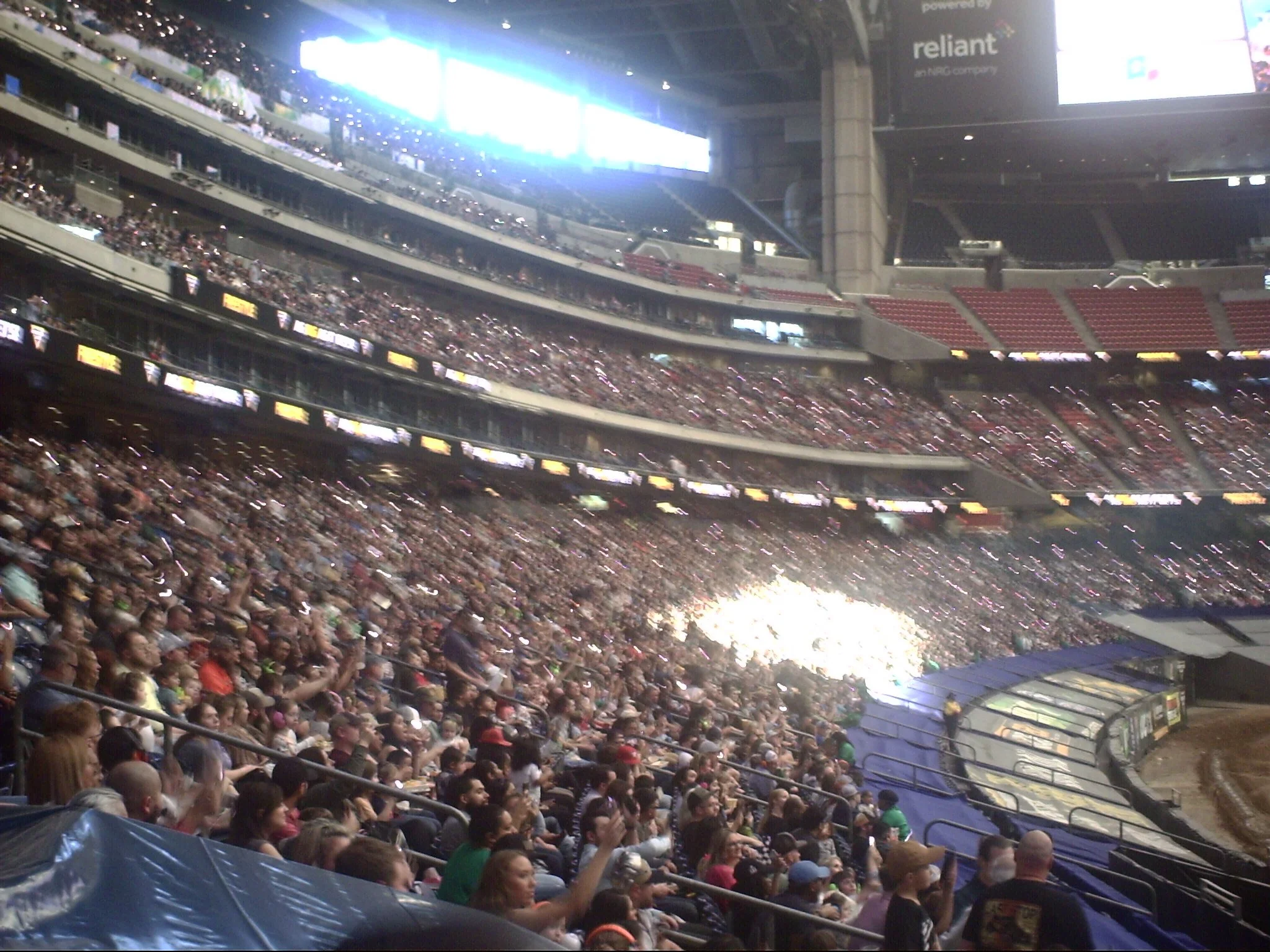 Crowd of people seated in a stadium with multiple levels, some in the foreground and others in the background, under a large roof with bright screens and advertisements.