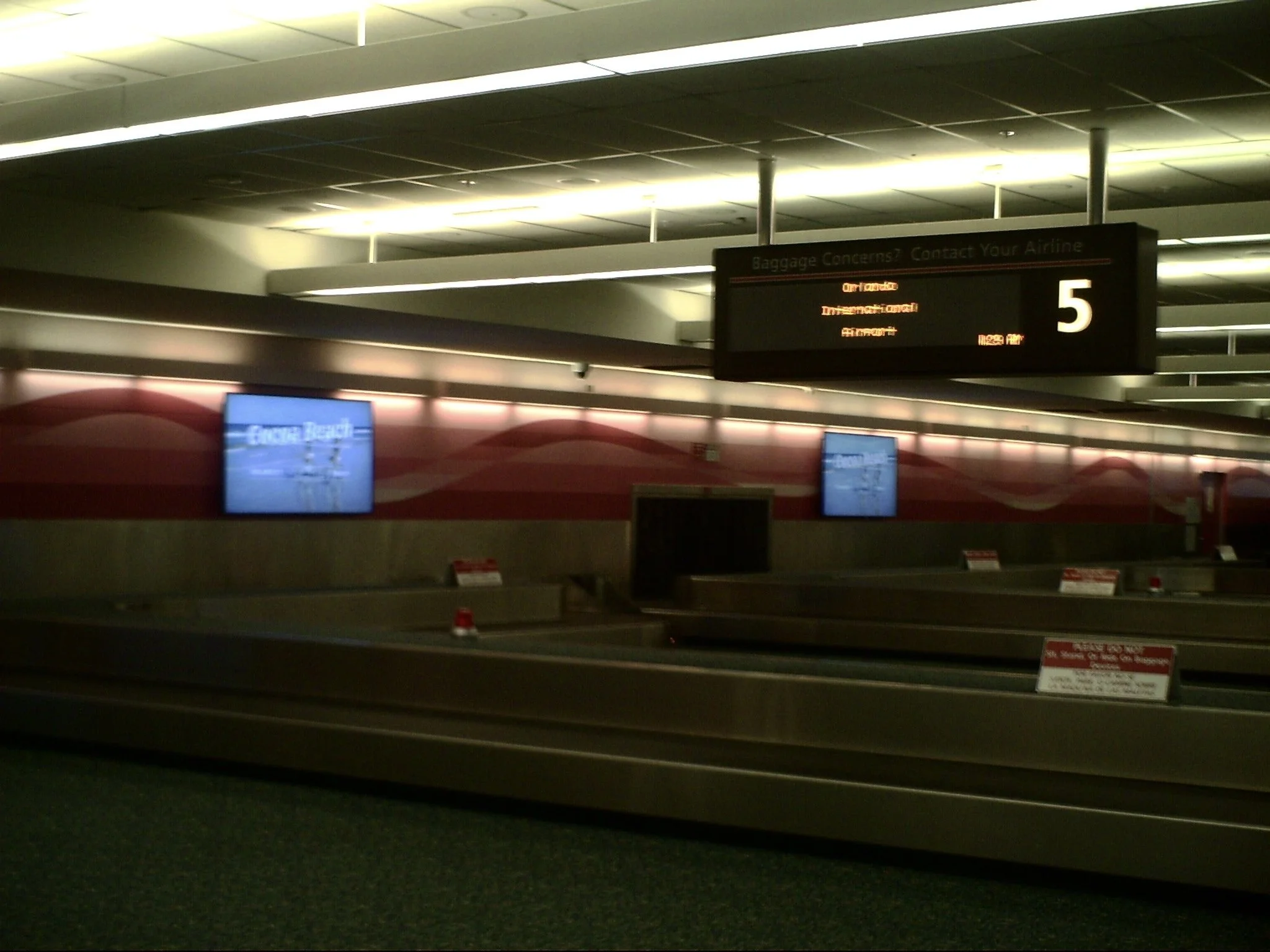 Airport baggage claim area with electronic display showing baggage information, conveyor belt in foreground, and advertising screens on the wall.